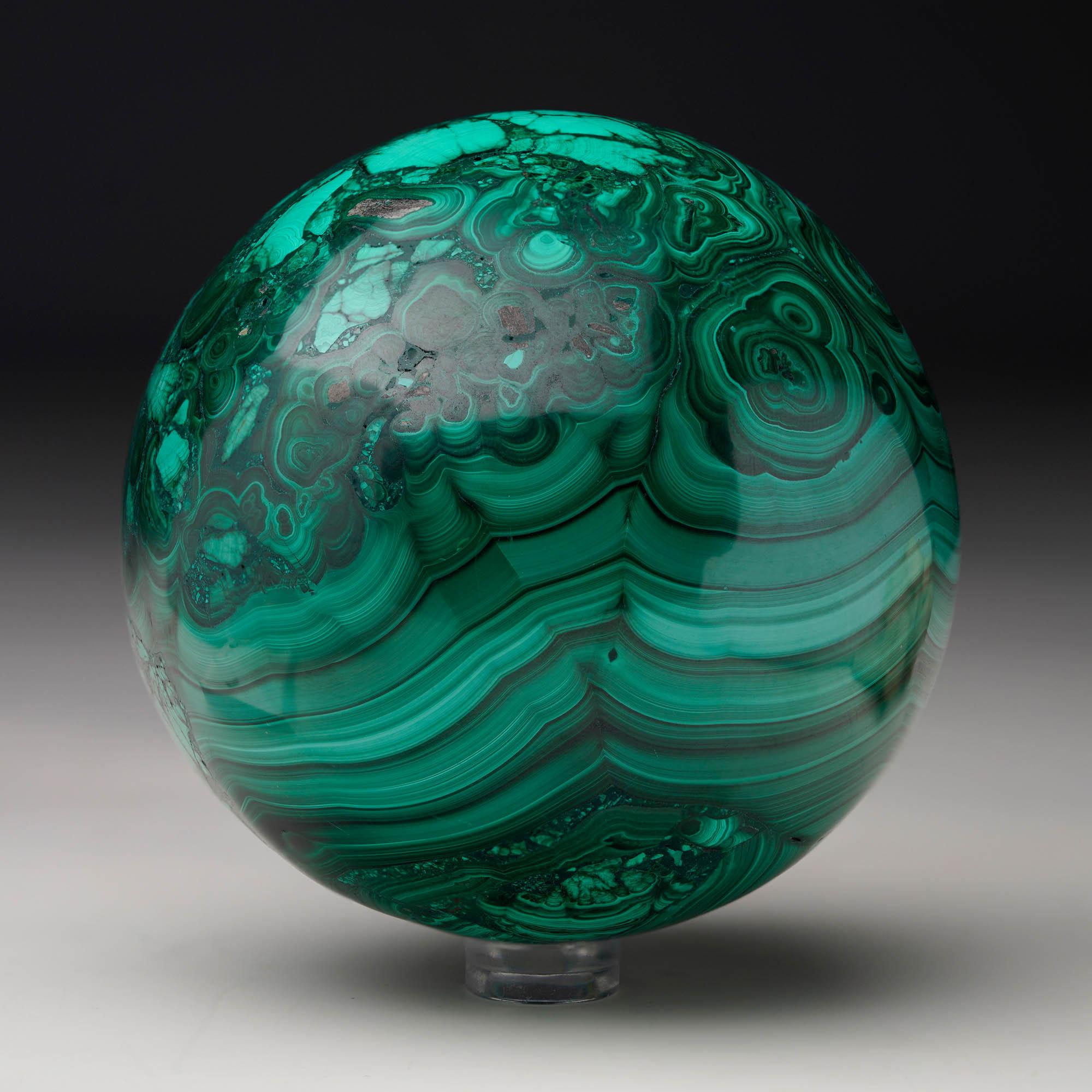 Crafted from solid, natural Malachite, this sizable hand-polished crystal sphere is a work of art: featuring a prominent, beautifully-defined banding, the Malachite's signature swirling green hues, contrasting bullseyes, and rich, natural luster