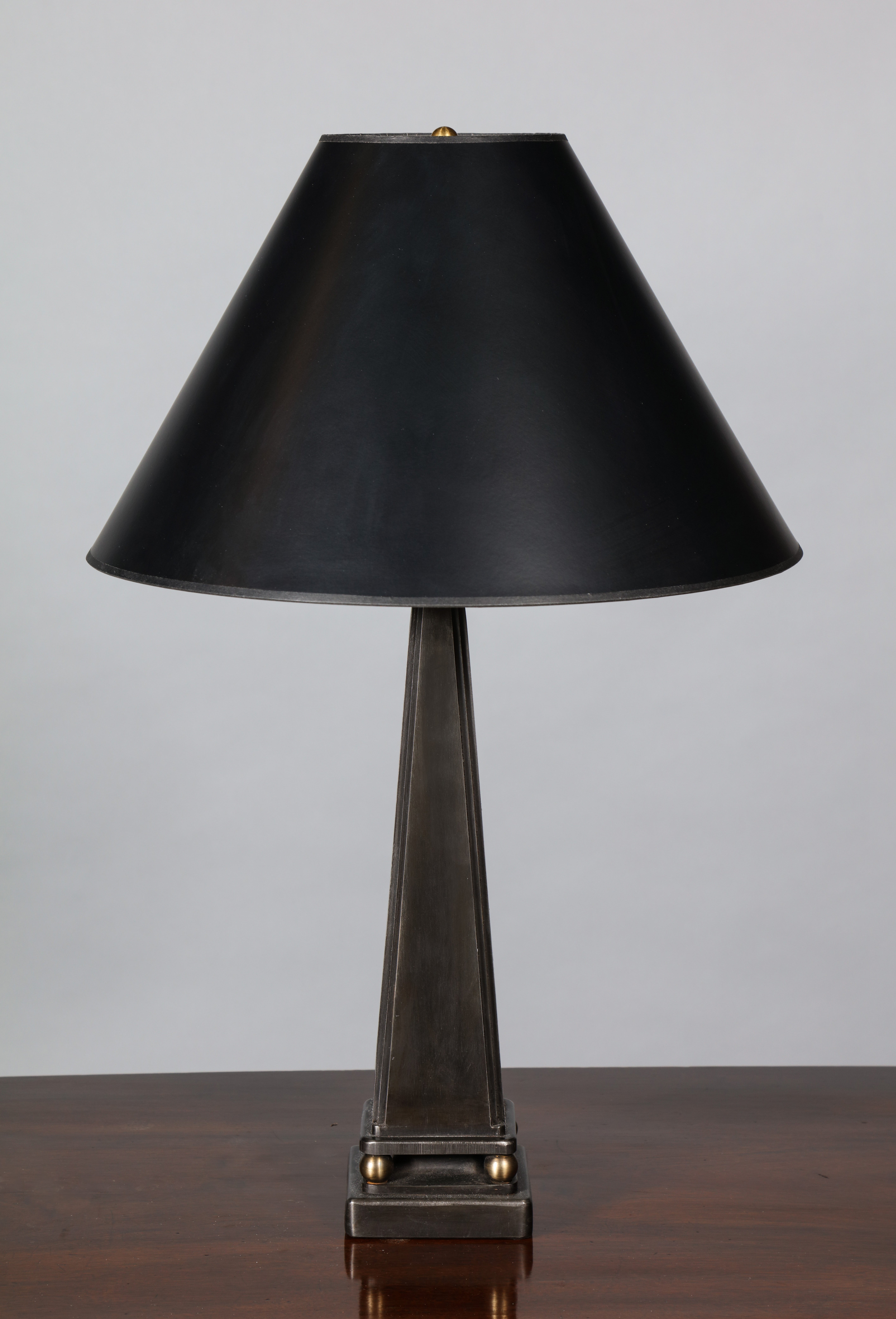 Lampe de table en acier fabriquée à la main, de forme pyramidale, reposant sur des pieds à boule et sur une plinthe carrée. Le design fait référence à l'esthétique néoclassique française des années 1930 à la fin des années 1940, avec ses cannelures