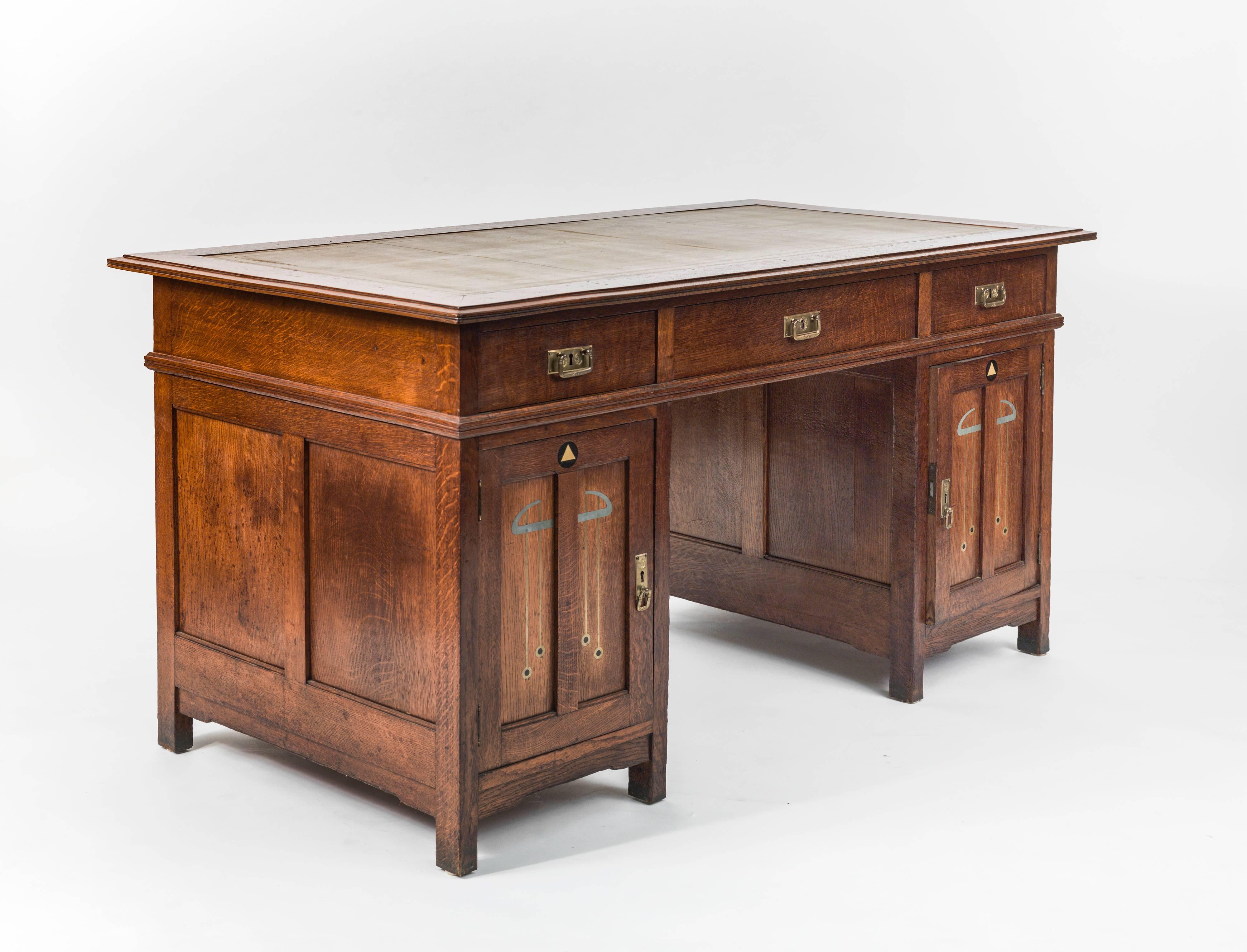 A Secessionist period oak pedestal desk, the apron fitted with three drawers; the pedestals inlaid front and back with period motifs in brass and pewter, the left pedestal arranged with vertical dividers for filing, the right pedestal fitted with