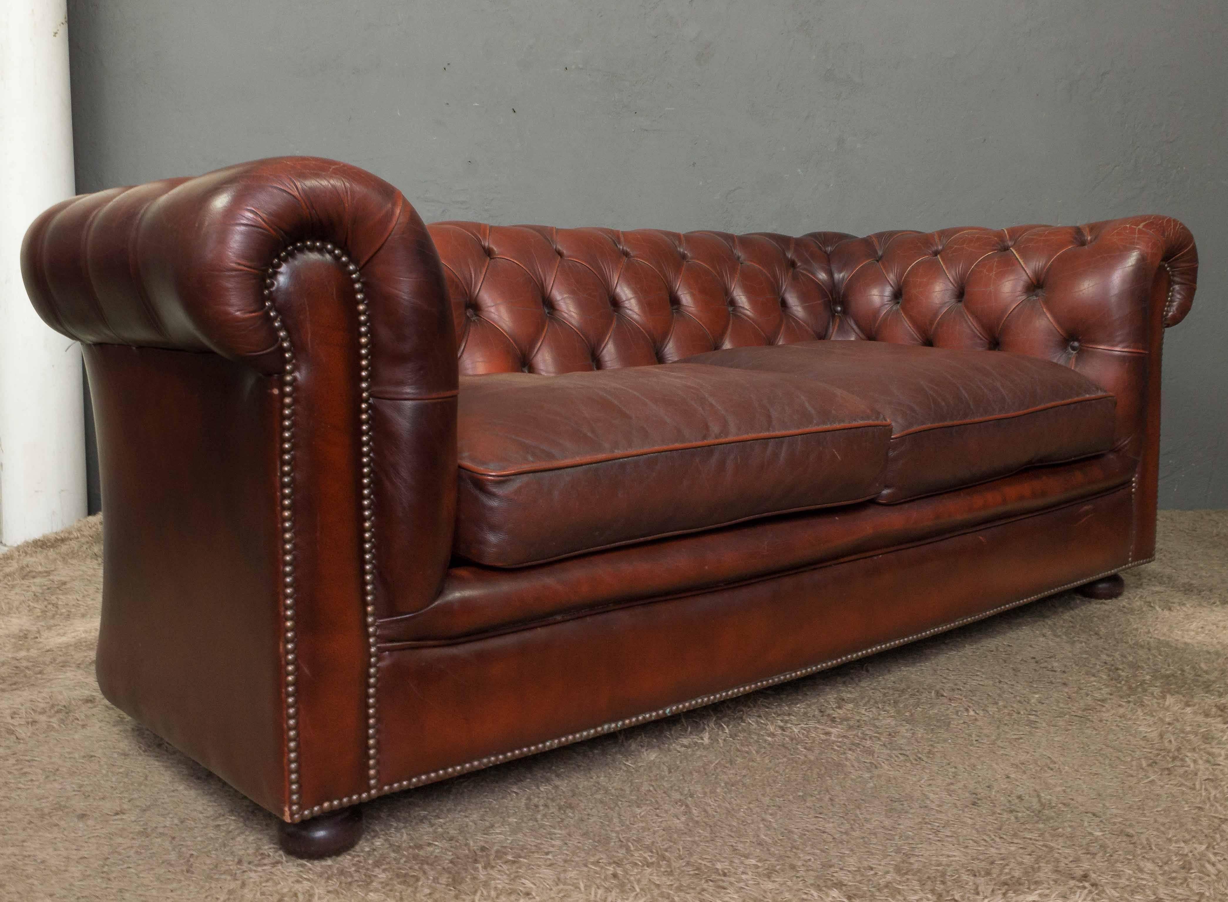 Classic red leather Chesterfield sofa with brass nailheads resting on wood feet. Leather has light cracking visible on arms.
 