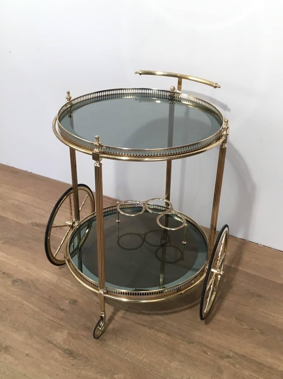 Neoclassical round brass bar cart with two blue and silver shelves with galleries and a three bottles holder. There are two large wheels on the sides and two smaller wheels on the front and on the back of the bar cart. The top shelf has four finials