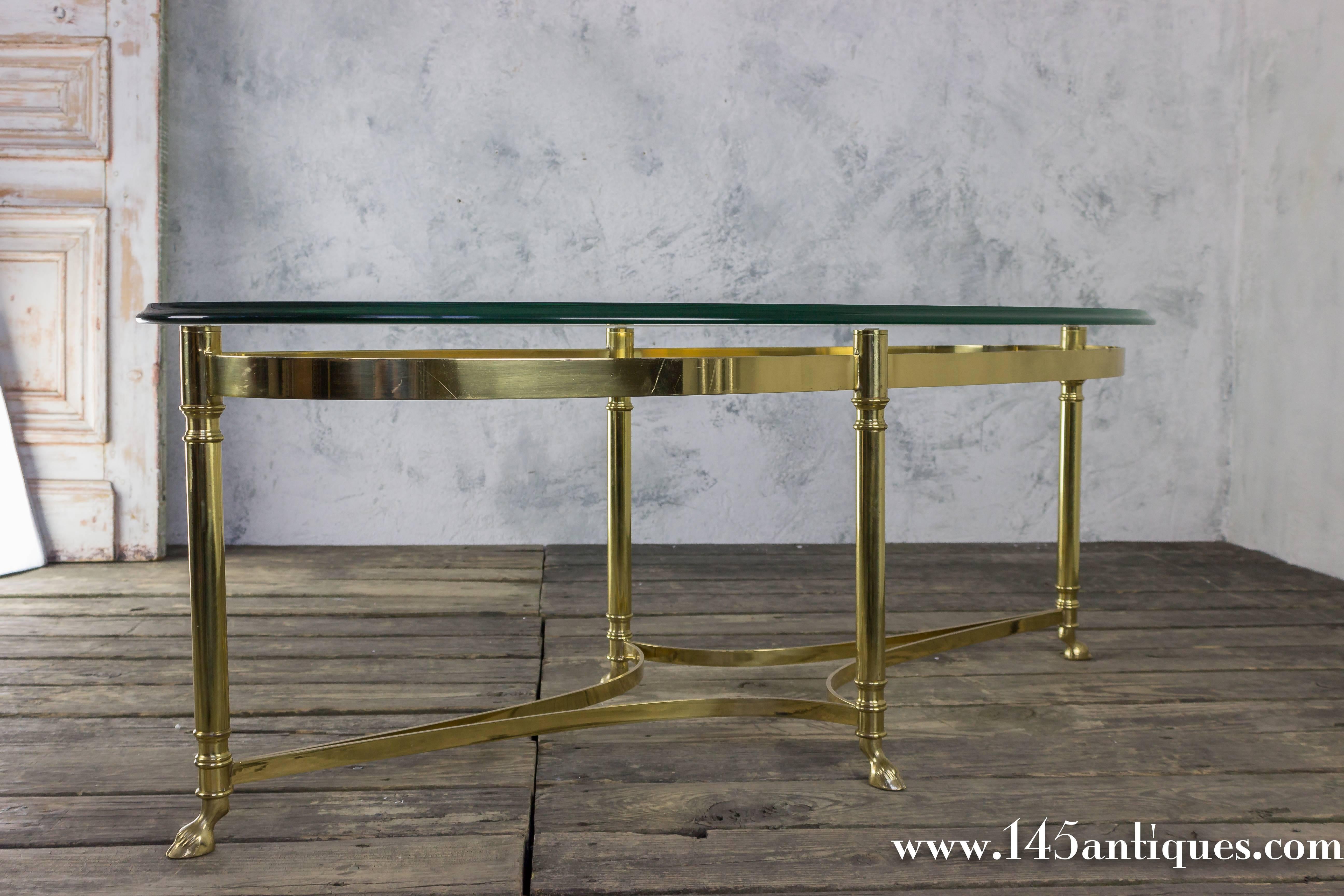 1940s oval shaped, brass frame coffee table with 'hoof foot' base. The glass surface has a rounded bevel with an elliptical shape.