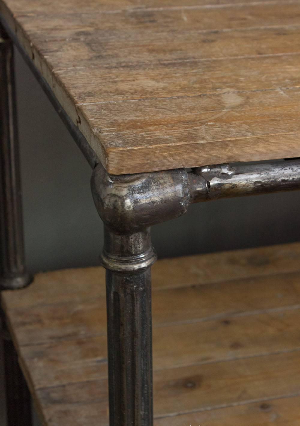 Questo antico tavolo da lavoro industriale è un'aggiunta notevole e funzionale a qualsiasi spazio abitativo, caratterizzato da gambe in ghisa scanalate in stile neoclassico che catturano l'attenzione. I due ripiani in legno forniscono un ampio