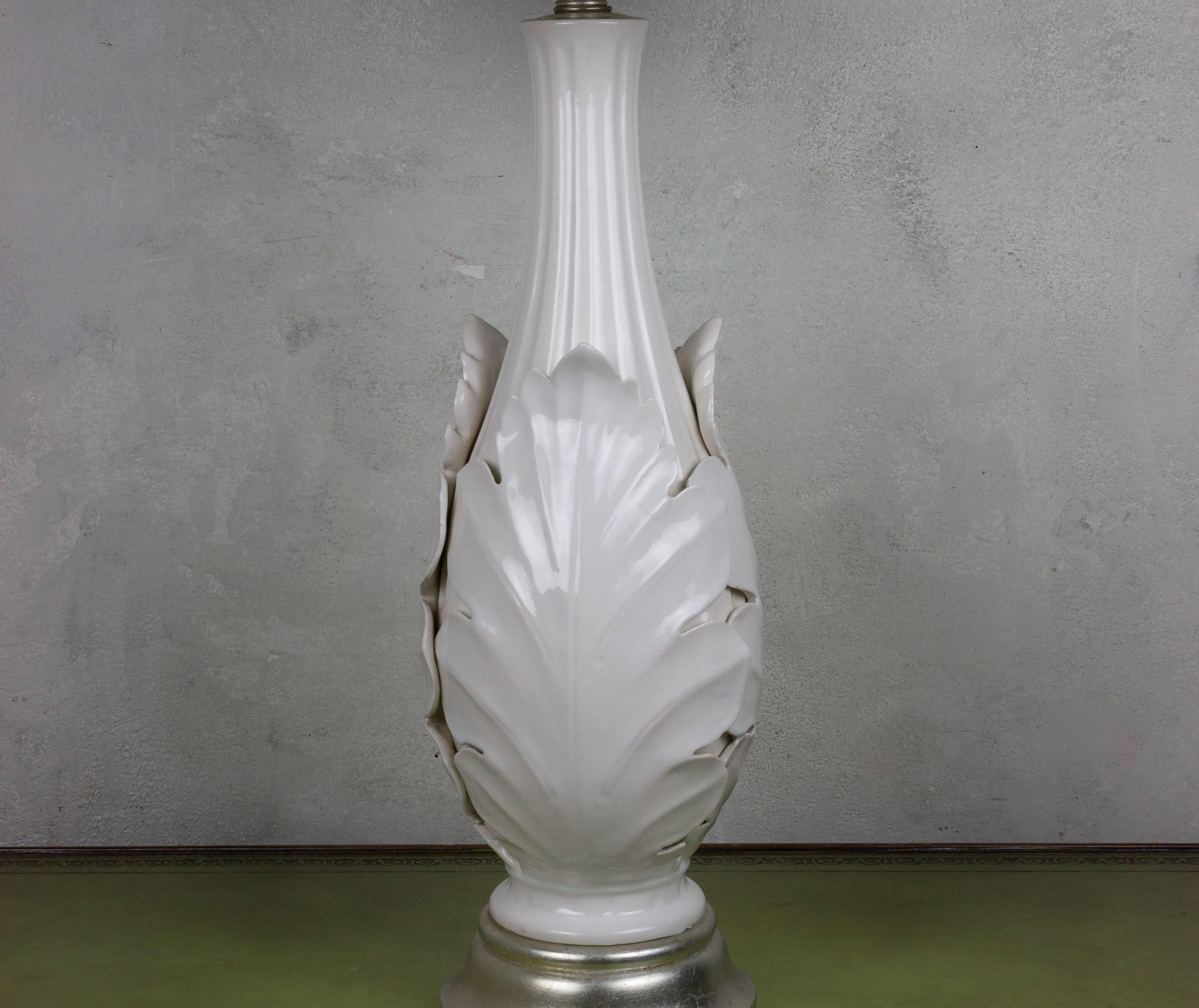 This elegant American 1940s white ceramic lamp embodies timeless sophistication. It features graceful stylized leaves, delicately encasing the stem, evoking a sense of natural beauty. Resting on a painted silver wood base, this lamp possesses a