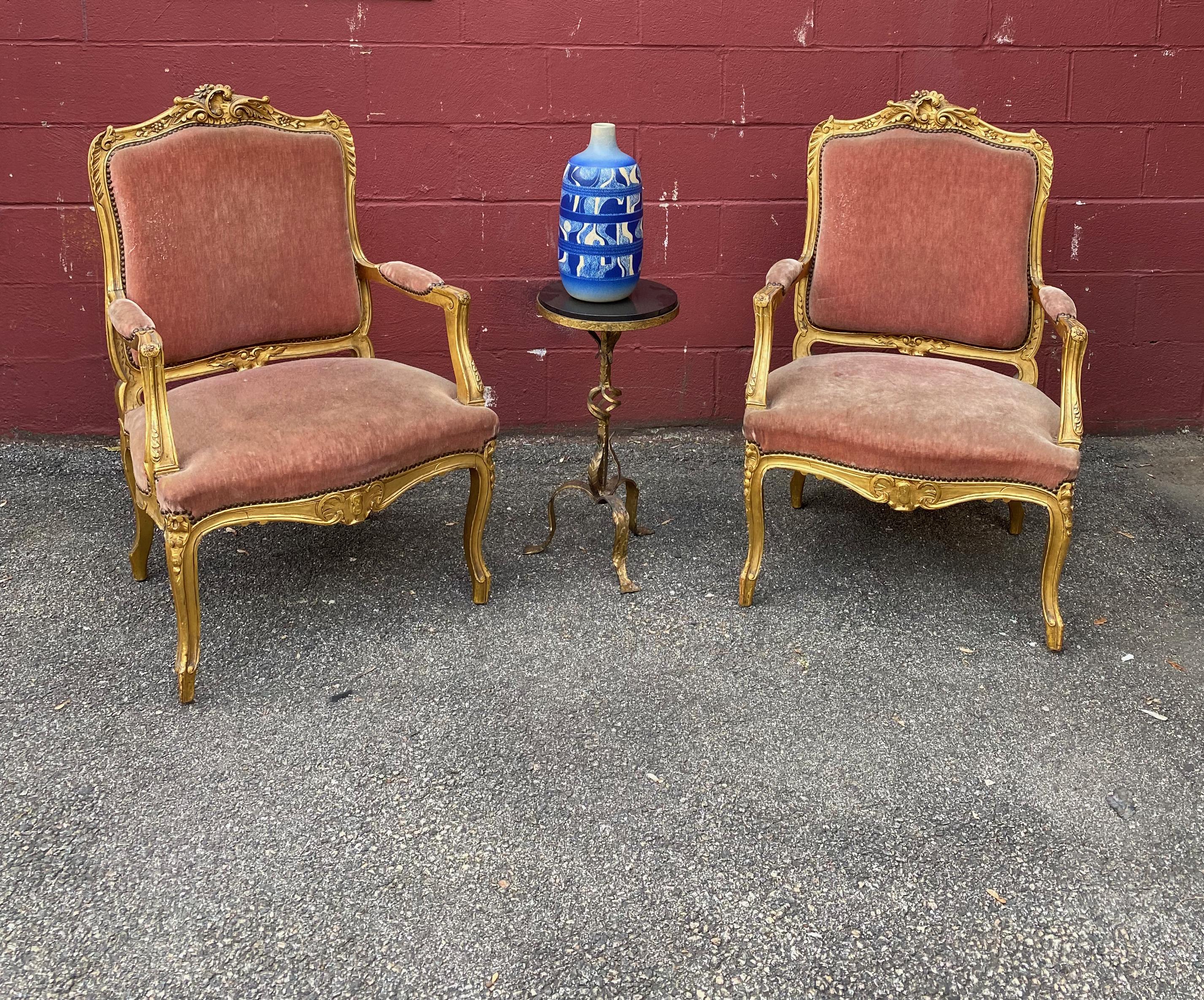 Pair of French late 19th century Louis XV style armchairs in a gilt finish with hand carved decorations. The faded distressed velvet is a deep salmon color. 
Fair vintage condition. Fades and stains on fabric, and the arms can use tightening.
