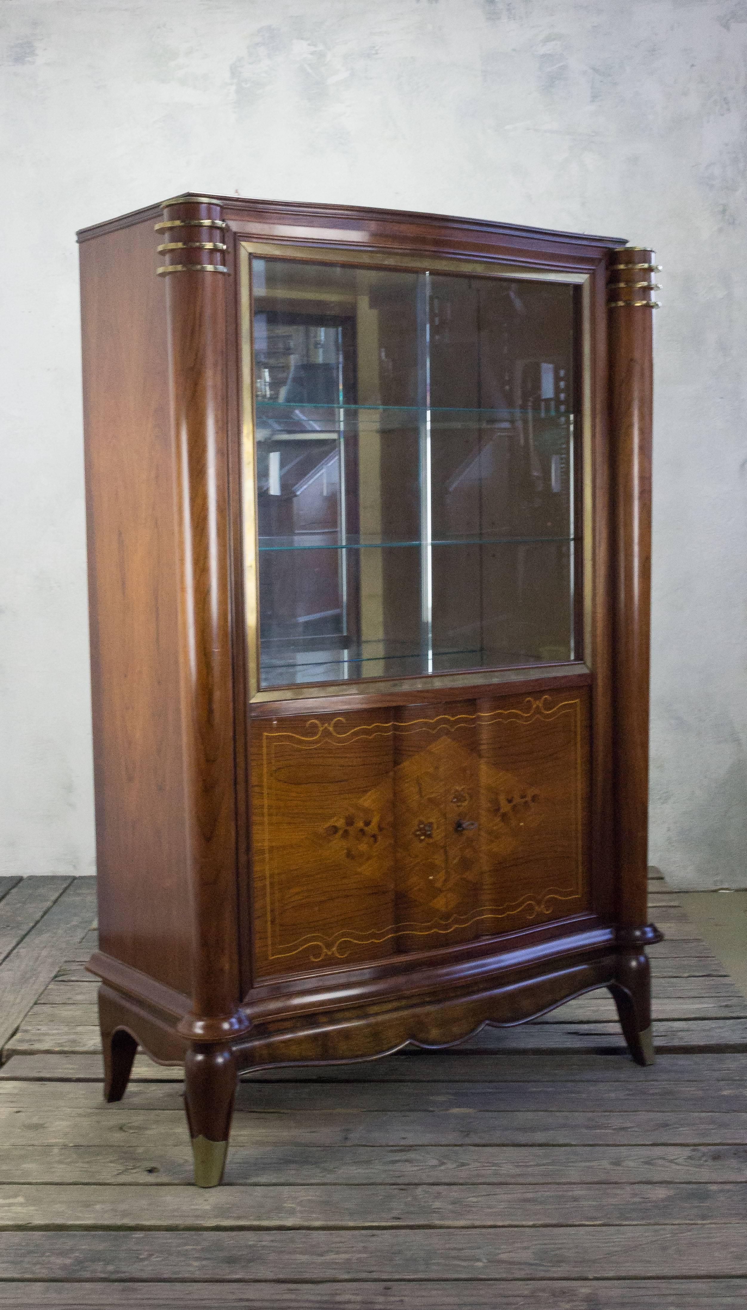 This French Rosewood display cabinet with inlays and brass sabot feet hails from the 1940s and is a true masterpiece of design in the style of Jules Leleu. The sliding glass doors elegantly protect the mirrored bottom and glass shelves, giving
