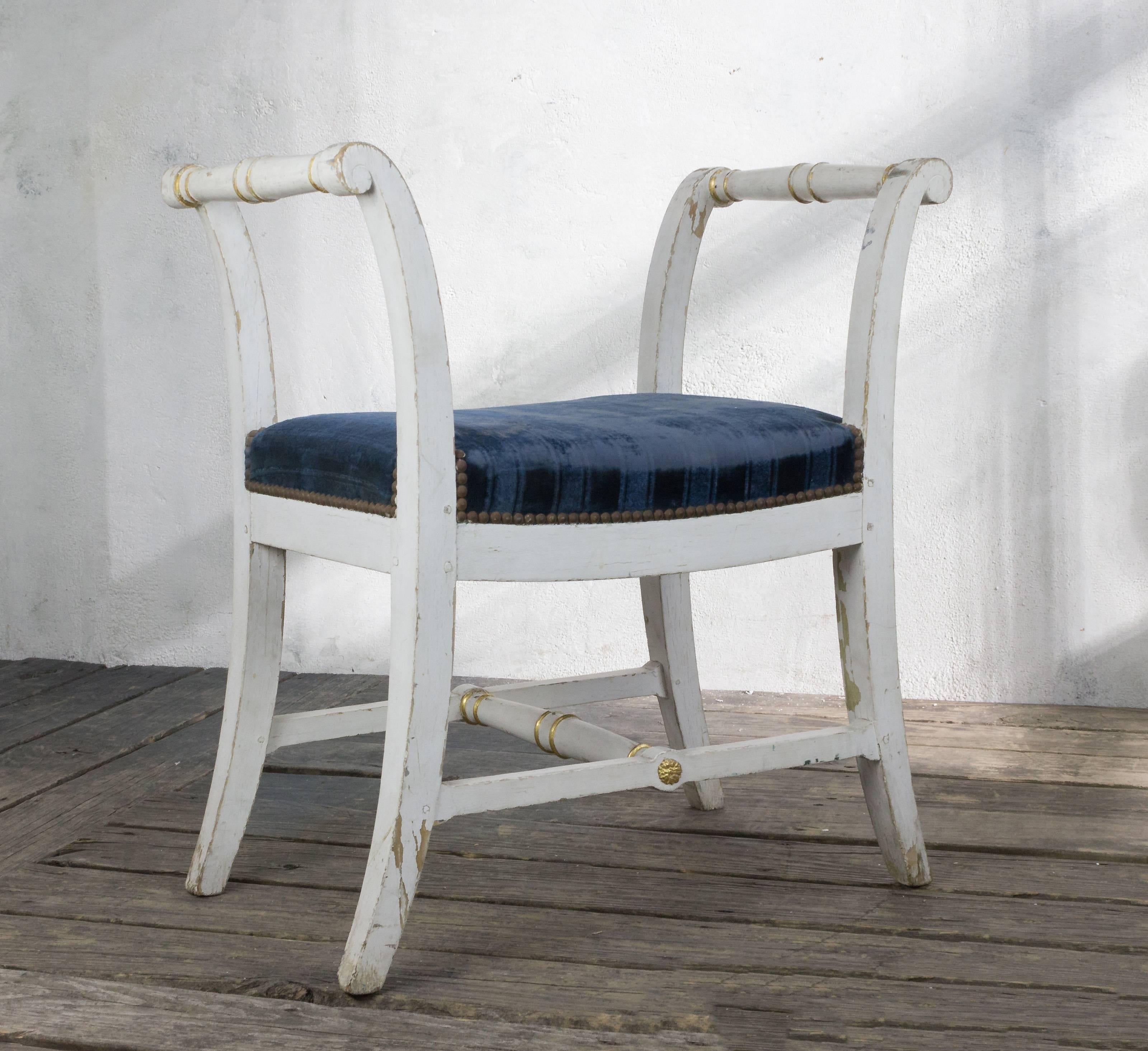A small Empire style bench upholstered in blue velvet. Add a touch of classic beauty and elegance to any space with this small Empire style bench. Boasting the original white painted finish with gilt highlights, this piece is upholstered in a