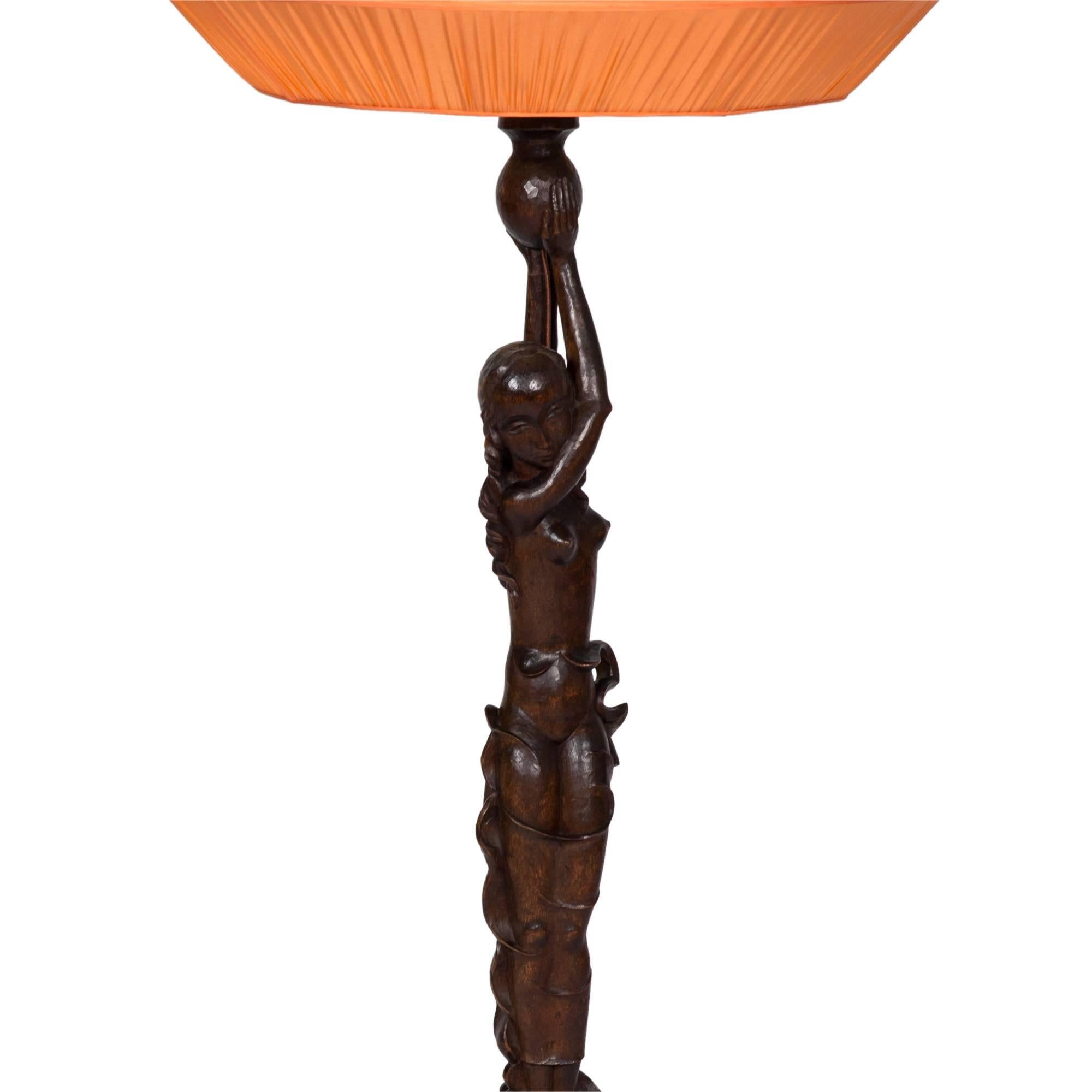 Carved-wood table lamp with three-bulb socket, in the shape of woman with custom, coral-color pleated silk shade. German, circa 1920. Measures: 45 in. overall height, 13 in. base diameter; 10 in. shade height, 20 in. shade diameter at widest point.