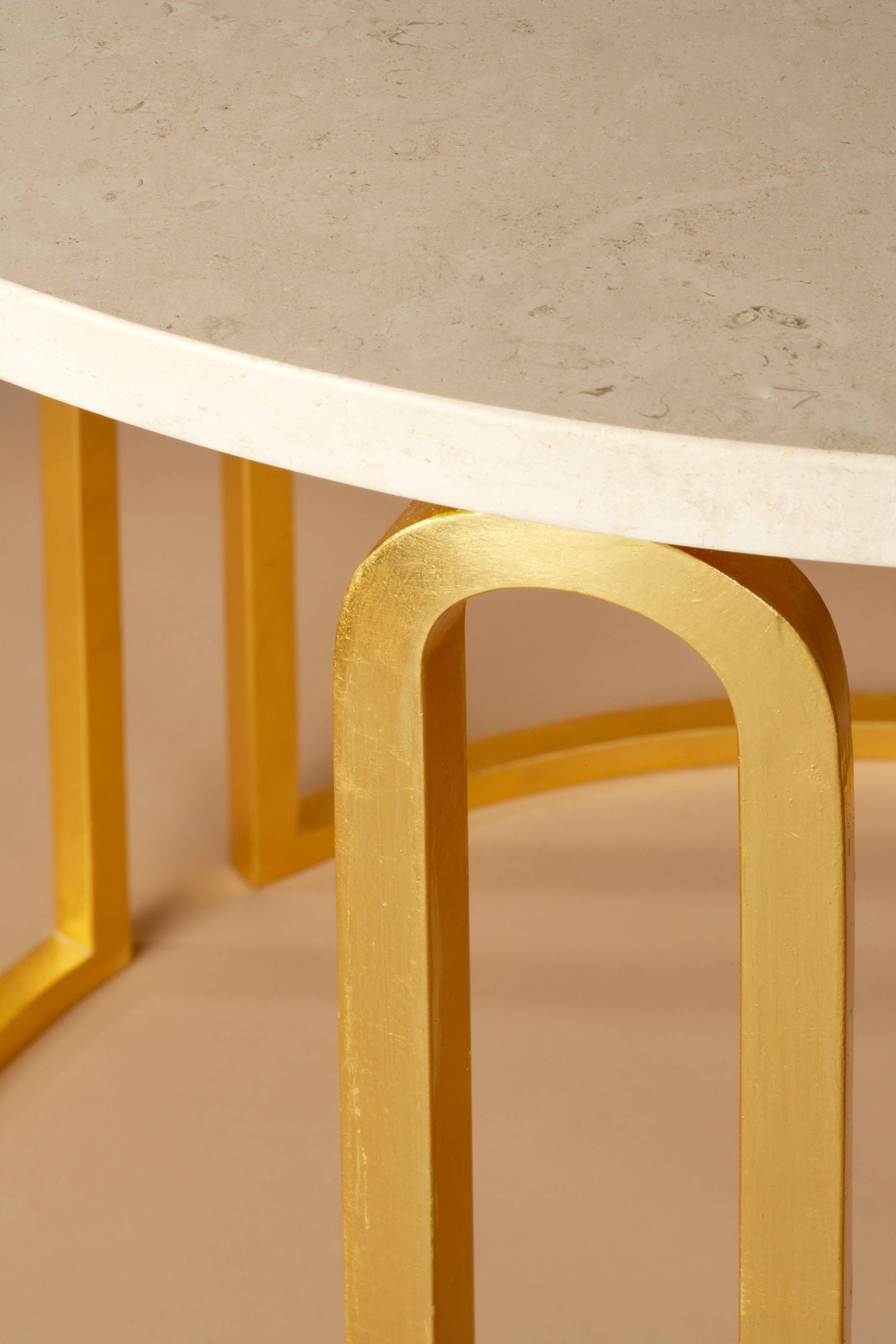 La surface lisse et soyeuse en travertin de Rhodes est soutenue par une base recouverte de feuilles d'or 24 carats, qui est forgée en trois arcs parfaitement proportionnés à partir d'une seule pièce d'acier. Cette table exquise est un parfait