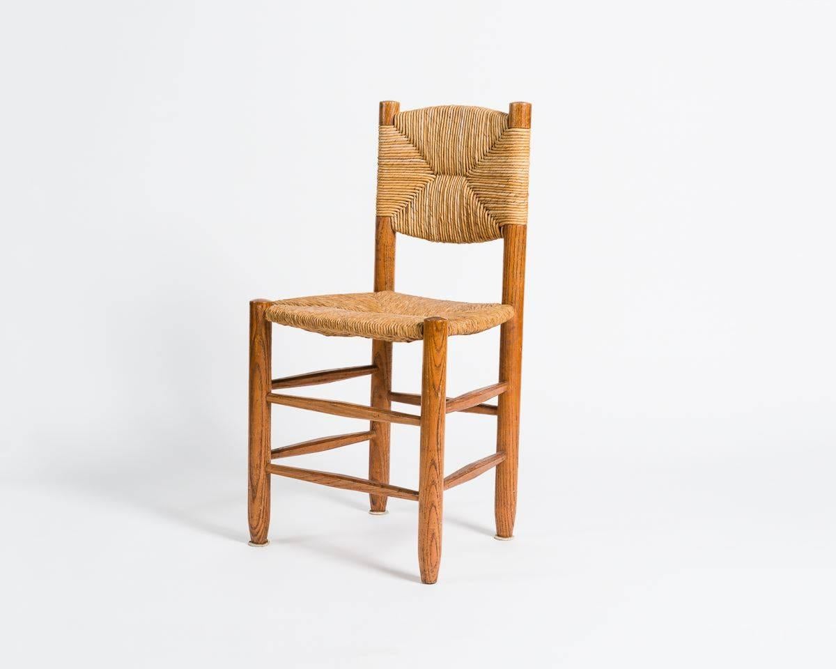 A pair of dining chairs of wood and straw in one of the quintessential Mid-Century designs of their creator, the famous French furniture maker Charlotte Perriand. 

These chairs are named Dordogne, a reference to a river located in Correze region,