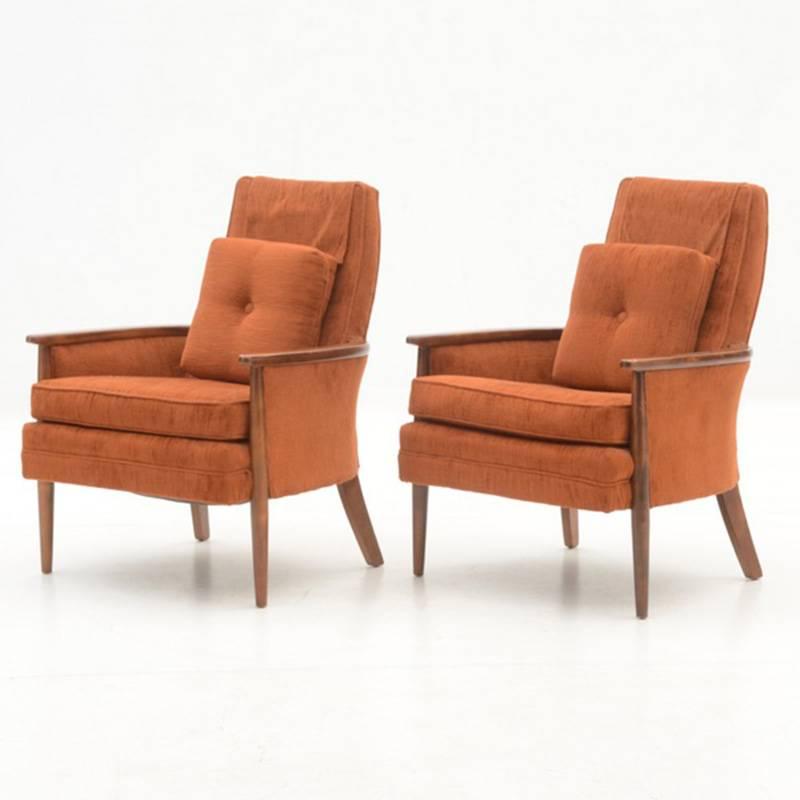 A pair of circa 1970s vintage armchairs, upholstered in burnt orange fabric with hardwood frames. Each has a dust cover to the top and includes a throw pillow.