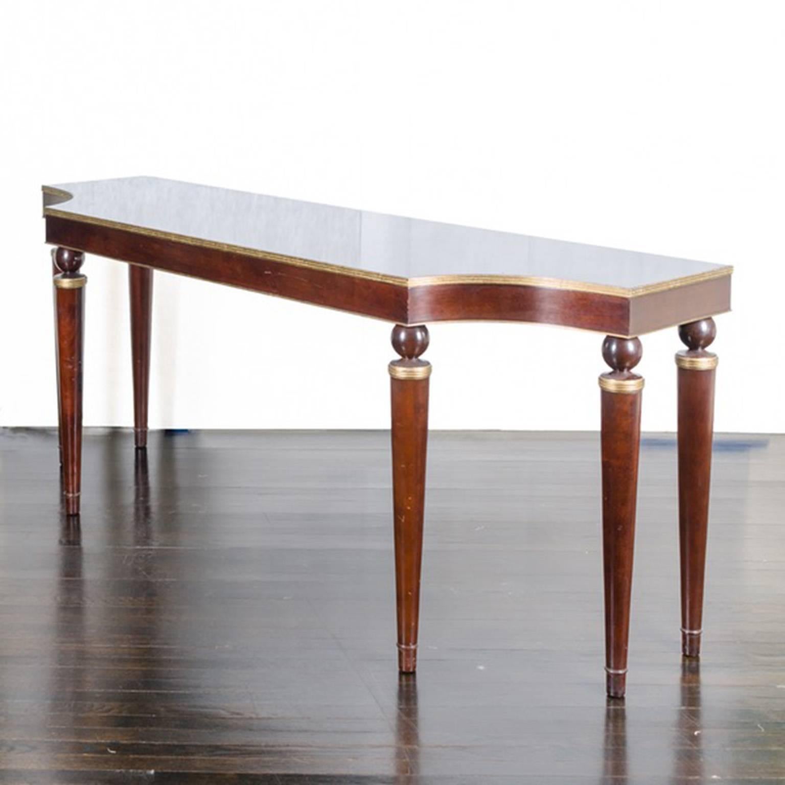 A contemporary mahogany console table with gold tone accents. The frame features a rectangular top with incurvate front corners and gold tone trim above a conforming apron. It rises on six tapered, cylindrical legs with ball capitals over gold tone