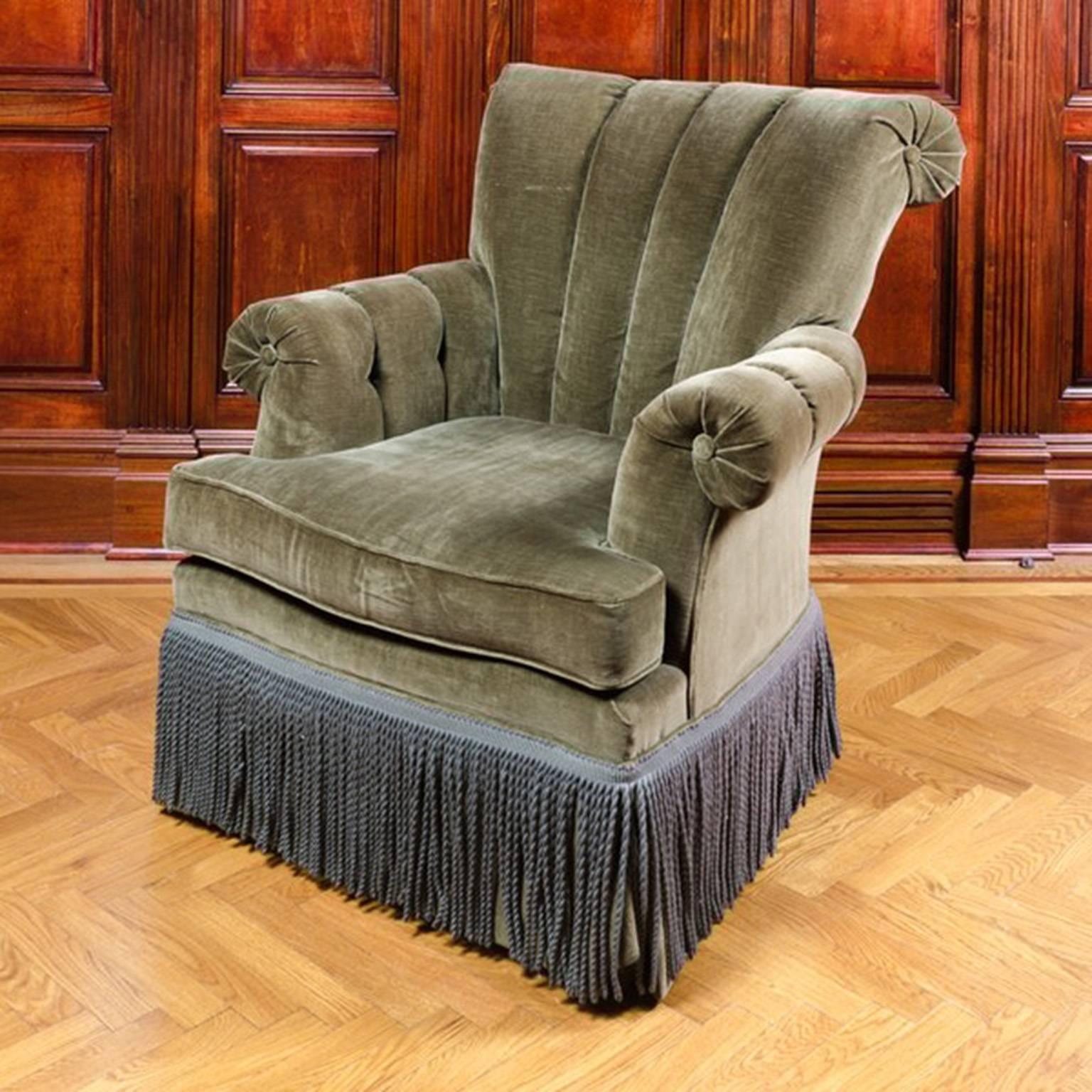 An armchair upholstered in olive green velvet. This chair features a rolled channel back and arms. It rests upon squared wooden legs and is embellished with a blue-gray fringe around the skirt.