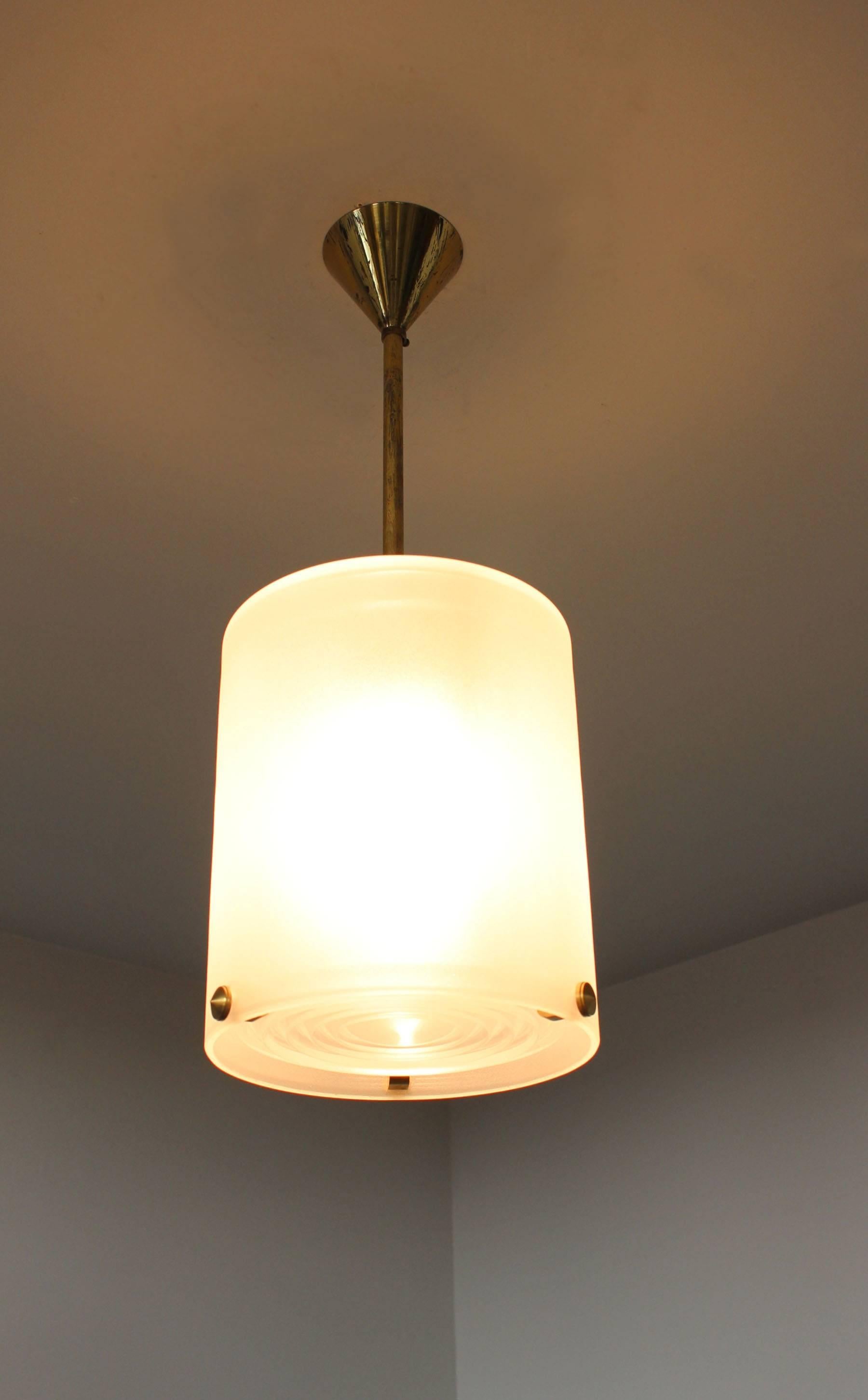 Un raffinato pendente Art Déco francese di Jean Perzel con un paralume cilindrico in vetro smerigliato e tre borchie in bronzo che sostengono una lente in vetro prismatico; anche l'asta e il baldacchino sono in bronzo. 
