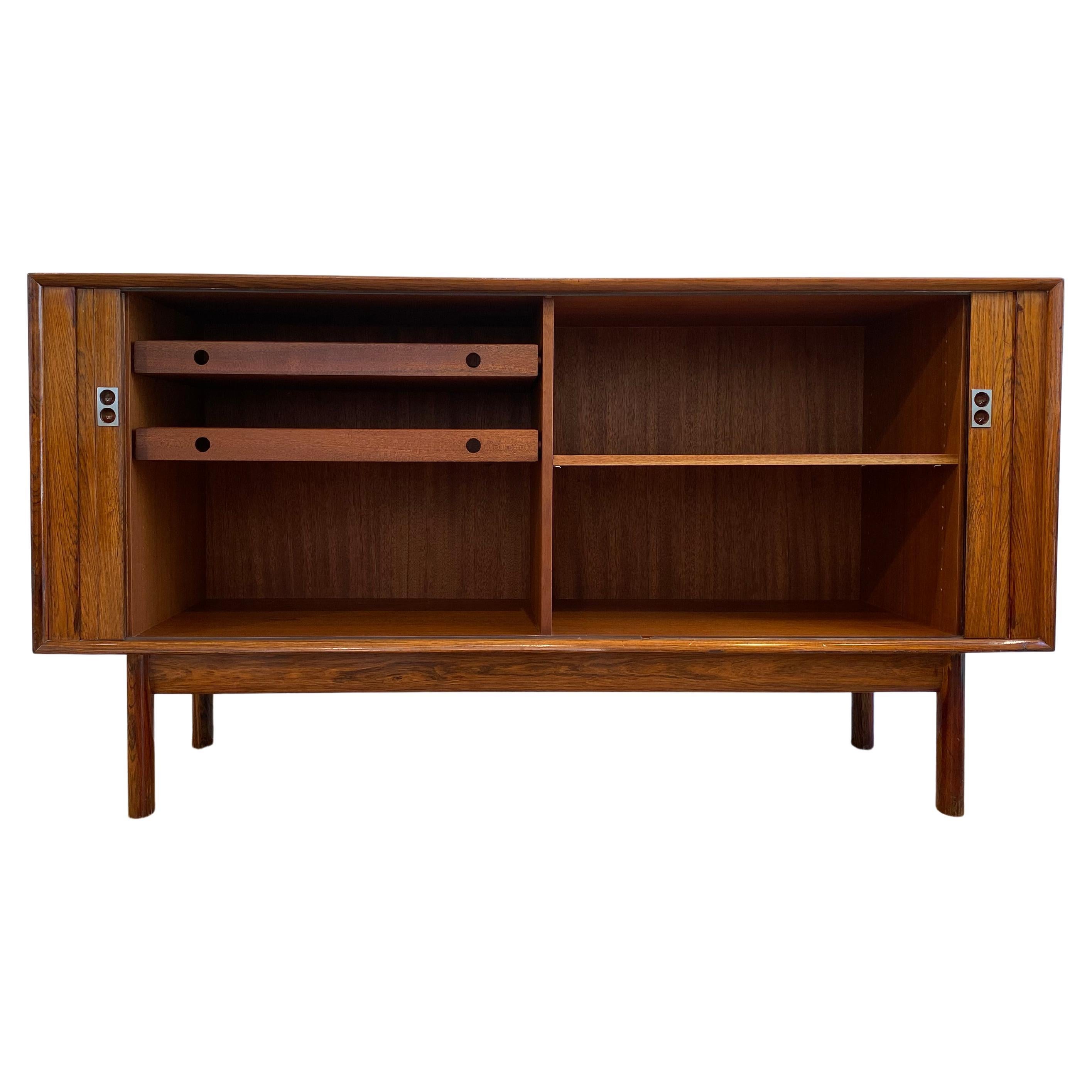Una raffinata credenza in palissandro di piccole dimensioni con una splendida patina invecchiata, disegnata da Arne Vodder per Sibast. Le sue ante scorrevoli a tamburo, le delicate maniglie in metallo e gli eleganti cassetti interni conferiscono una