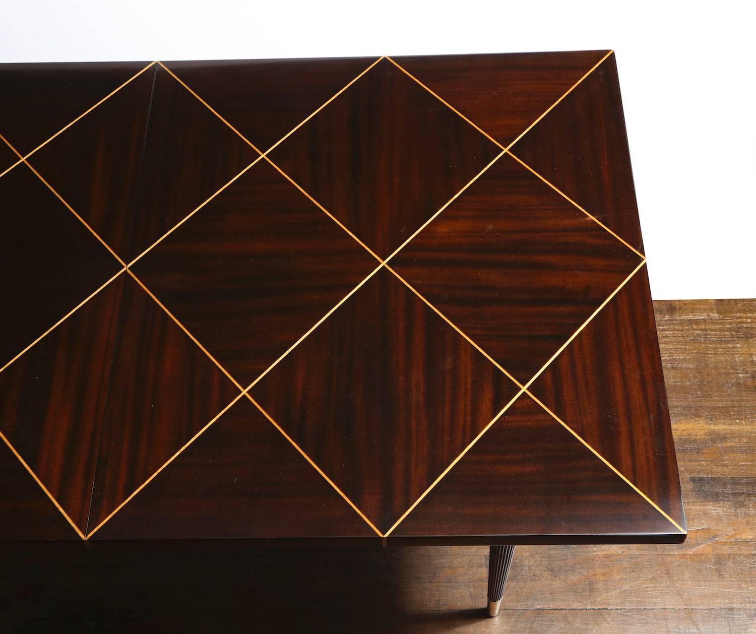 Dark mahogany table with veneered top in alternating grain and light holly inlays. Four fluted legs that taper and end in brass boots. Two extensions that measure 17