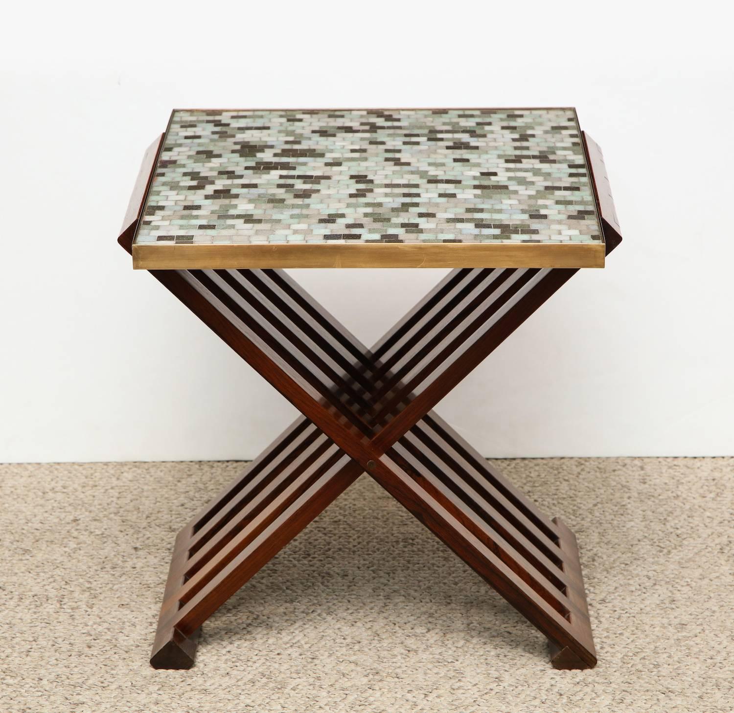 Side table #5425, by Edward Wormley for Dunbar.
Beautiful interlocking and folding wood base with highly figured graining. Brass-rimmed top with inset Murano glass tiles of pale blue, green, brown and white. Green Dunbar tag attached to underside