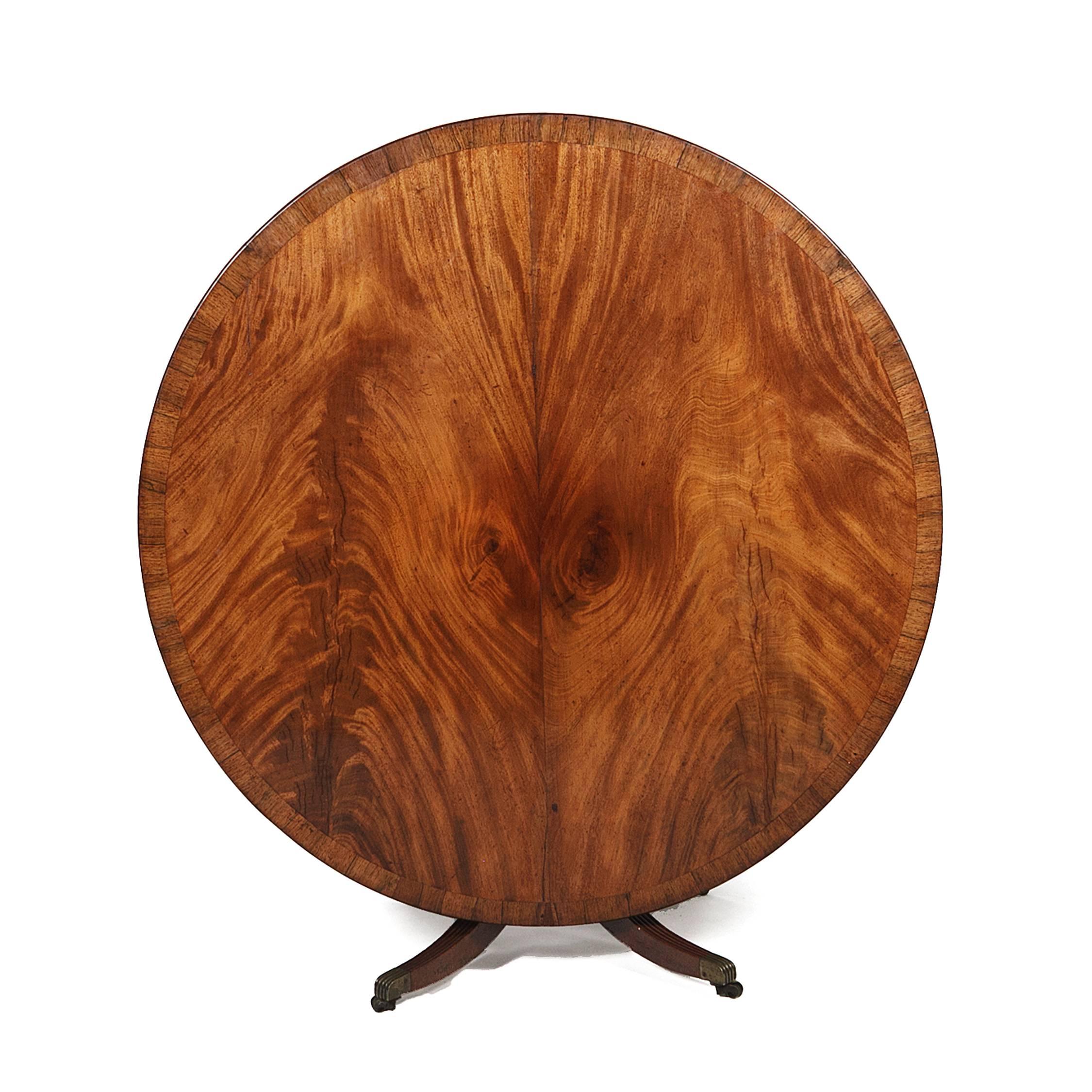 This circular mahogany center table, with a rich finish and a striking grain, is supported on a single turned pedestal with four reeded, outsplayed legs, and with brass toe caps and casters.