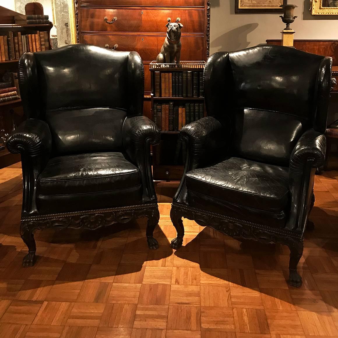 This pair of 19th century black leather armchairs is distinguished by the Georgian-style shell and acanthus carving on the apron and cabriole legs of each chair, ending in stylized lion's paw feet. Each of these highly comfortable and generously