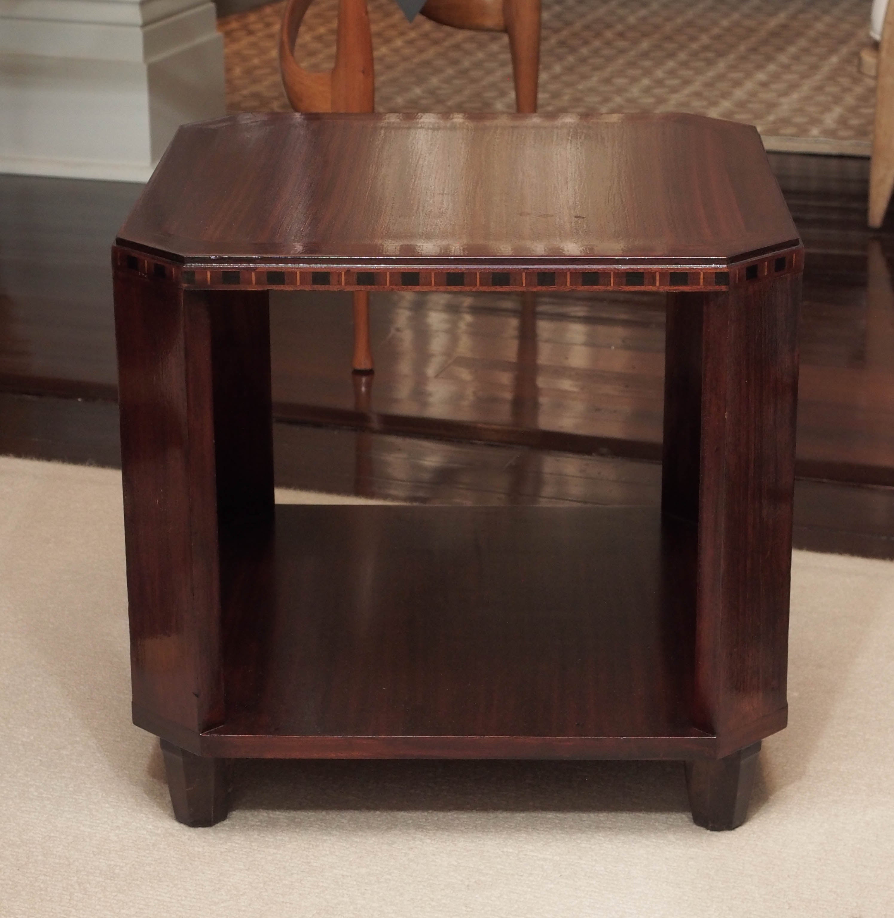 Two-tier side table in mahogany; canted corners; the top apron with a band of ebony and fruitwoods; hexagonal feet.