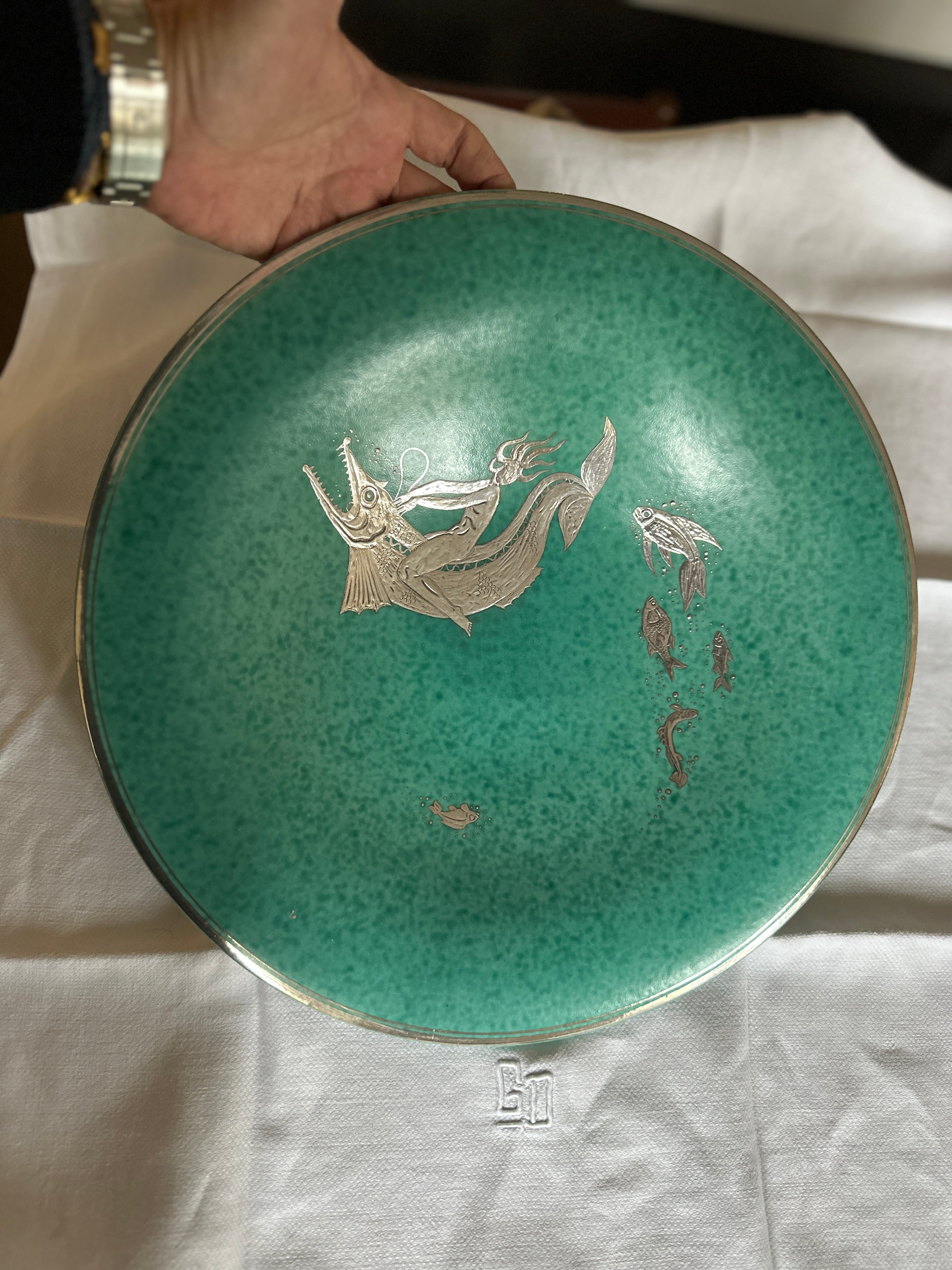 Très grand centre en grès émaillé  avec recouvrement d'argent Dimensions : 15 pouces de diamètre Cette belle pièce fait partie de la série ARGENTA que l'artiste a réalisée pour la manufacture de Gustavsberg en 1917 et qui lui a valu son premier