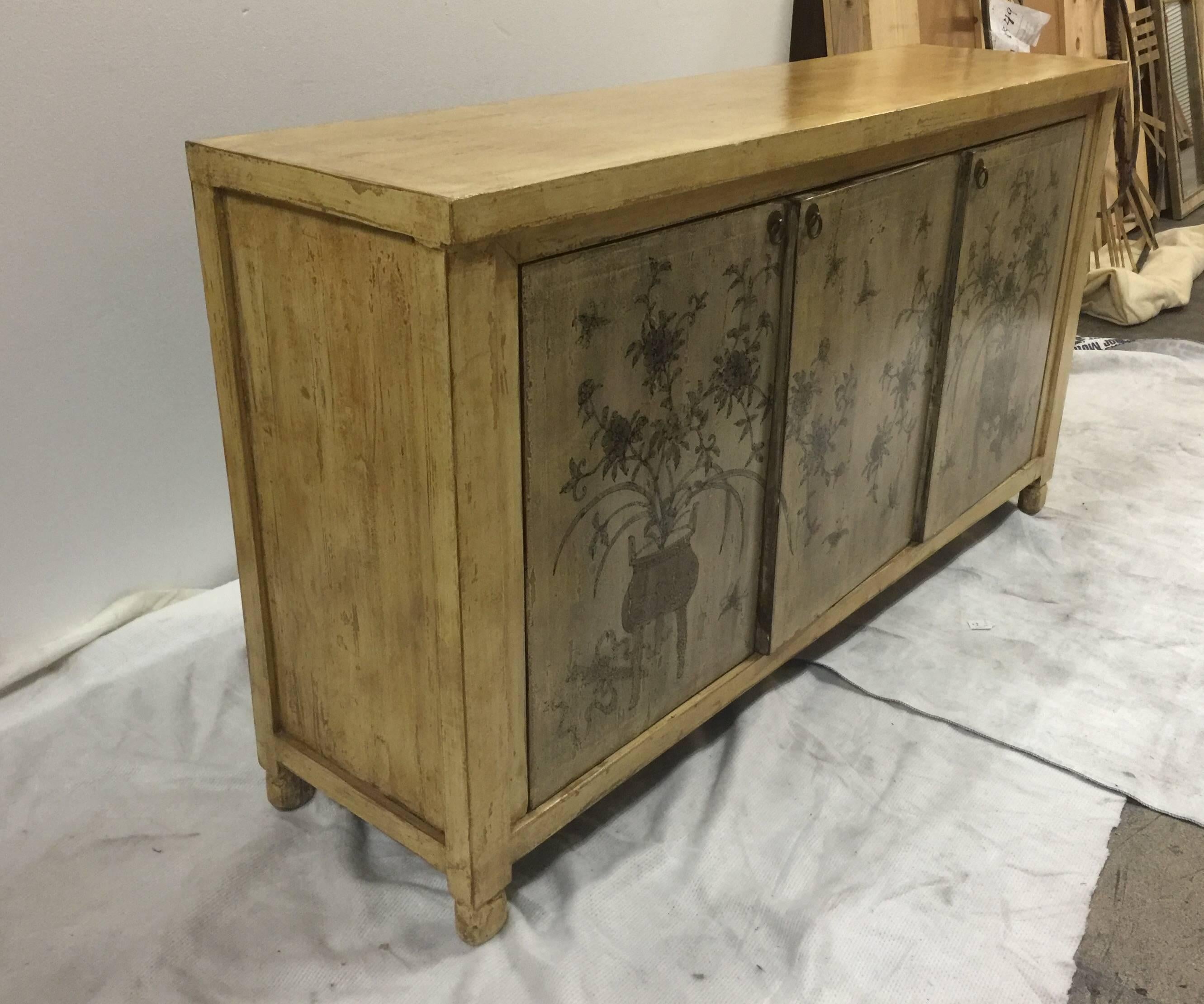 An Asian-style credenza or sideboard with three door fronts featuring a hand-painted floral design, USA, circa 1970. Three doors open to reveal adjustable interior shelves. Case made of blond wood with a mottled stained finish.