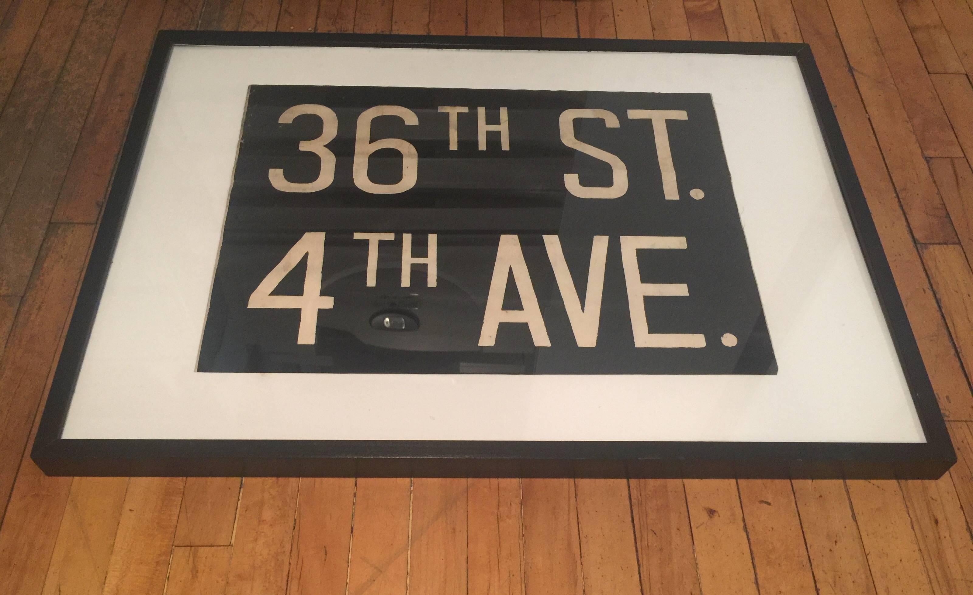 A segment of a vintage New York destination sign in black canvas with white text, circa 1950. Originally used on a subway (or bus) window sign on a roller mechanism. Newly mounted on an off-white linen matte, pleixglass and black wood