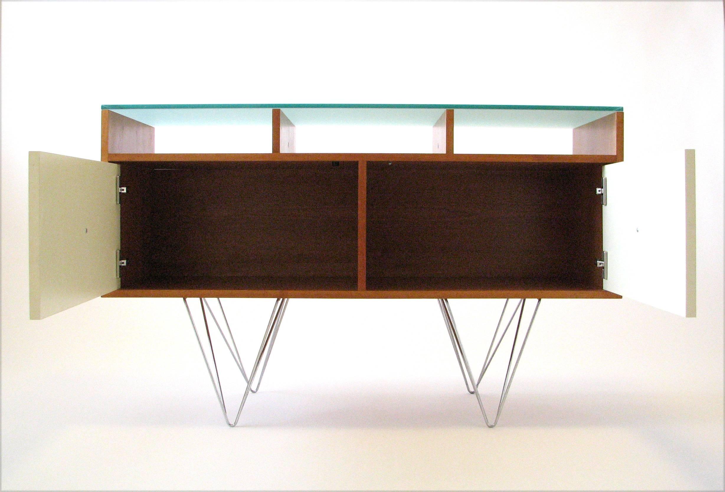 A cherrywood credenza or bar service cabinet with a frosted glass top over three open compartments, full-width storage behind two ivory melamine-wrapped doors, and chromed double hairpin legs. The finished cherry back makes it fully suitable for