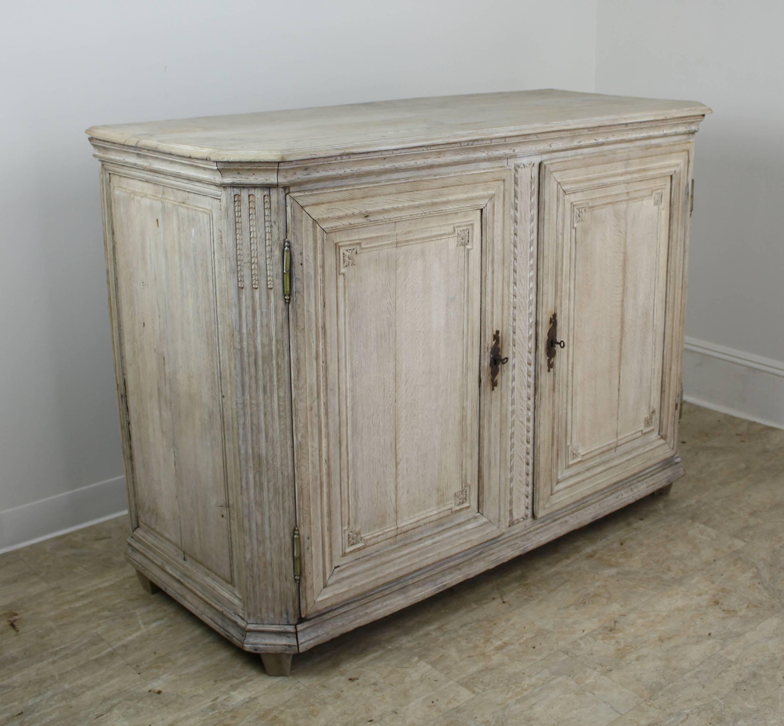 A stunning tall French antique buffet in bleached oak. The exterior is fancifully decorated with hand carvings, specifically in a floral motif at the corners of the four doors. Chamfered corners on the top and charming escutcheons add to the visual