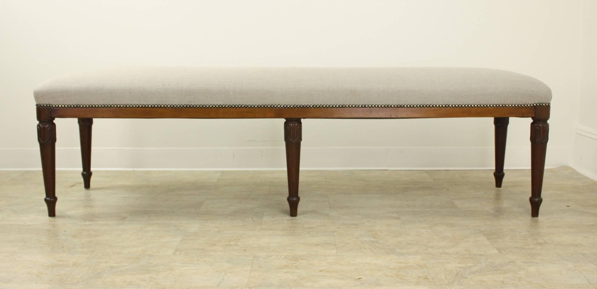 An elegant long stool newly covered in French beige linen, with brass decorative upholstery nails. Constructed with  mahogany legs c1840 that are well-carved and sturdy. Very good sitting-height, comfortable seating. Suitable as a long ottoman in