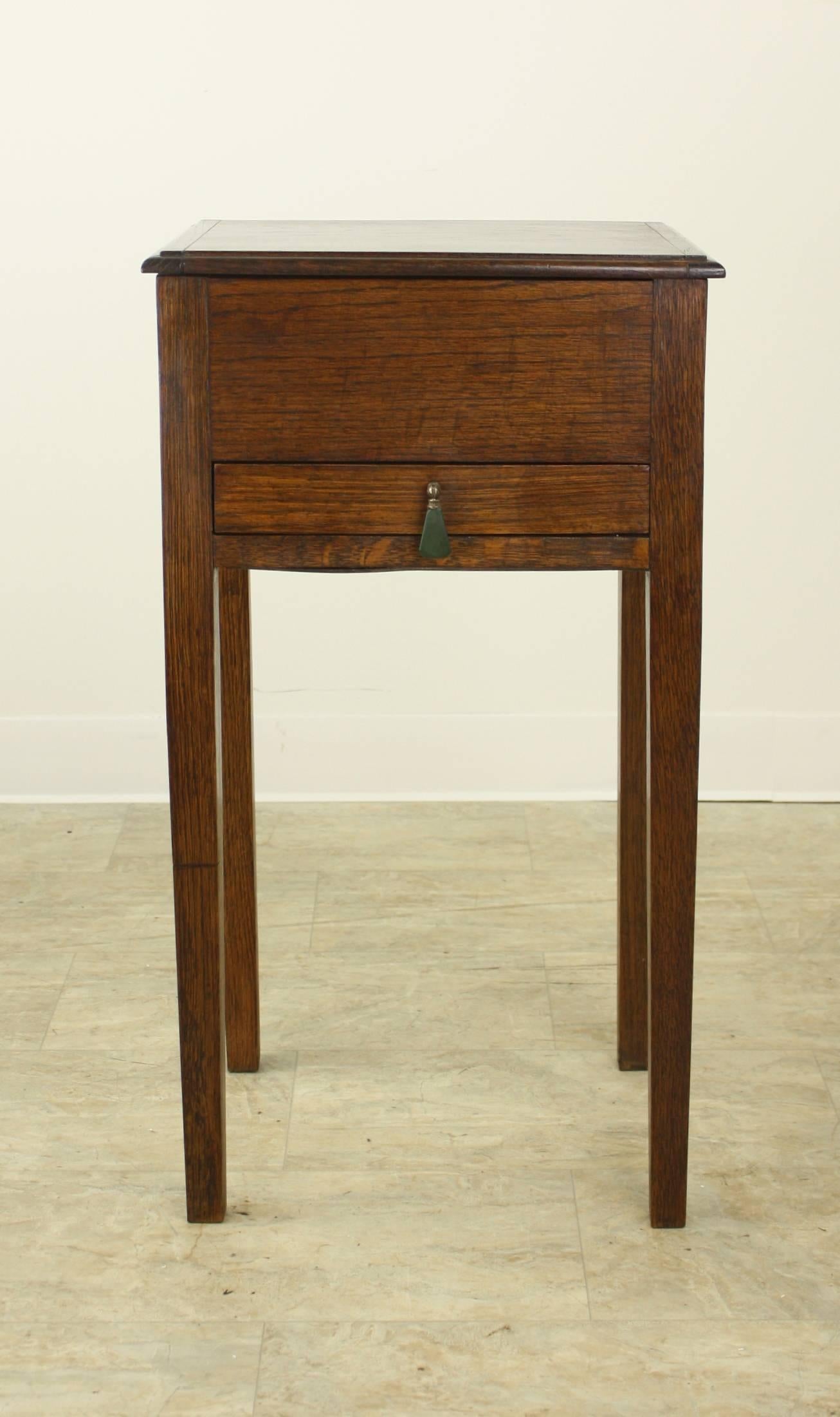 A small oak sewing box on long tapered legs makes a charming side table! Wonderful deep color and patina. The top flips open to a deep storage bin, and there is a small sliding drawer with a decorative green teardrop pull. Perfect for as a lamp or