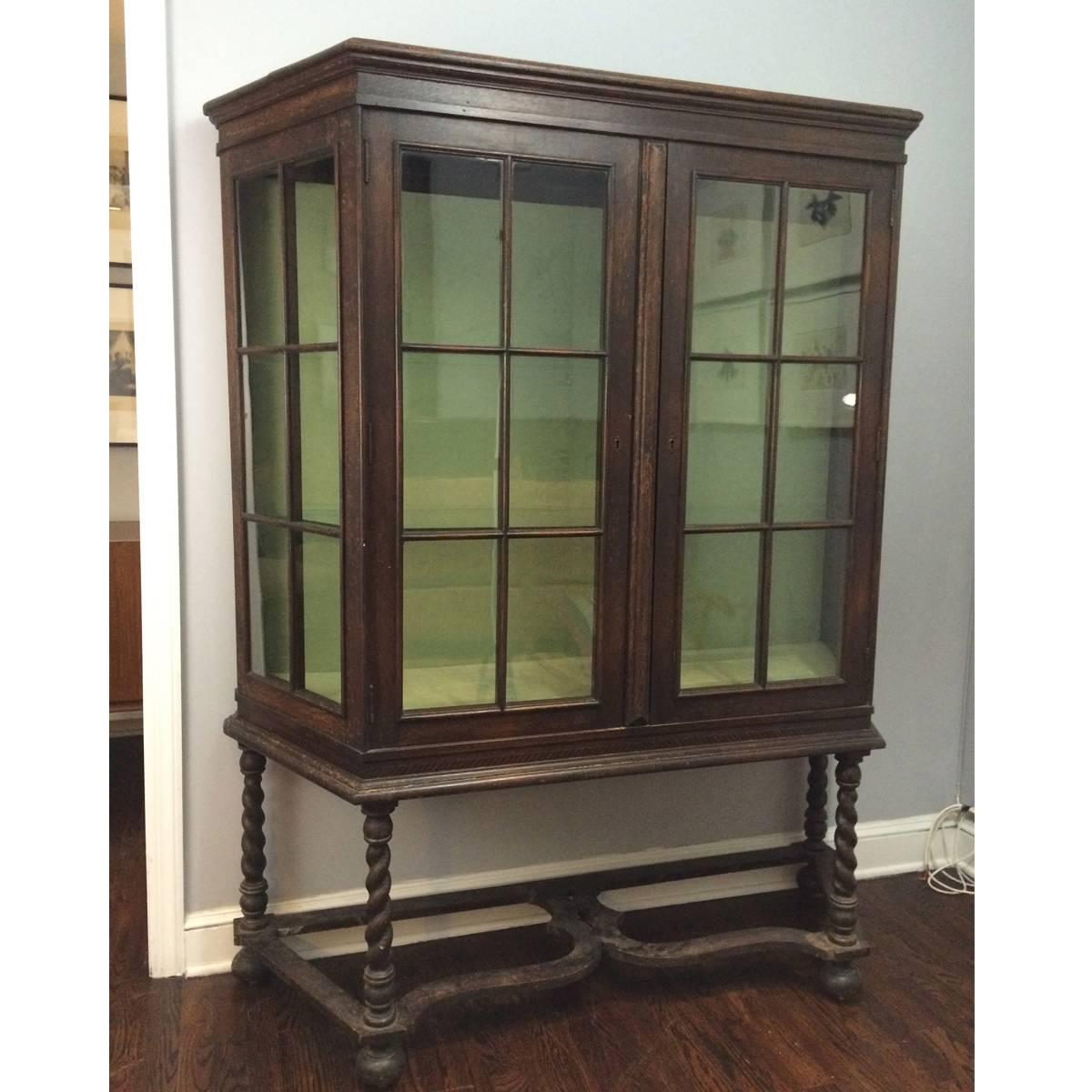 Dark oak curio or China cabinet in the Jacobean Revival style comprised of a felt-lined glass case with two doors and two glass shelves resting on a base of four barley twist legs connected by stretchers on bun feet. Cabinet is trapezoidal and