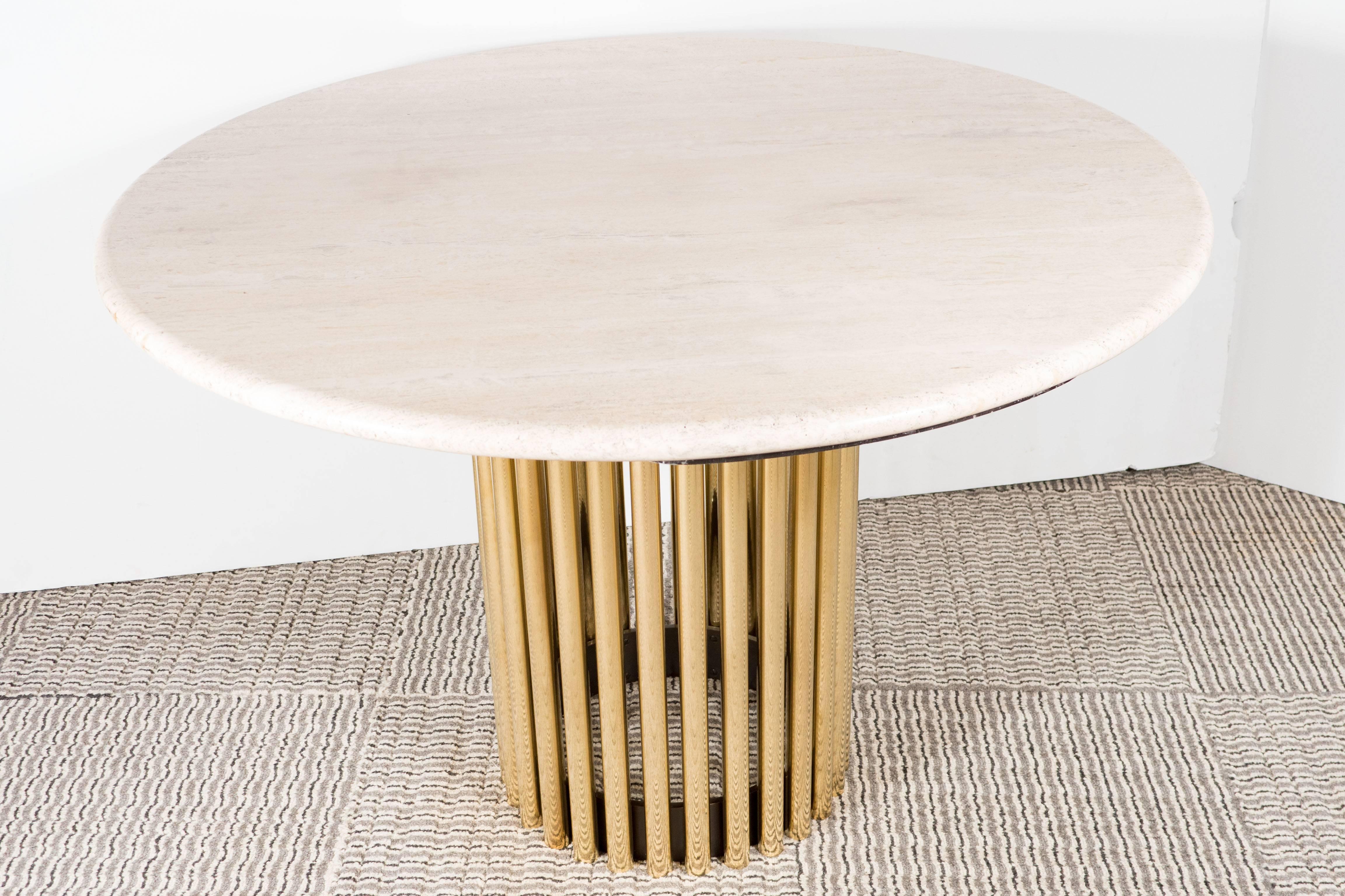 A Mid-Century Modernist dining or center table featuring a polished brass tubular base. The brass rods are connected by an enameled base ring. The base supports a polished, circular travertine top, which displays warm hues of cream and beige notes.