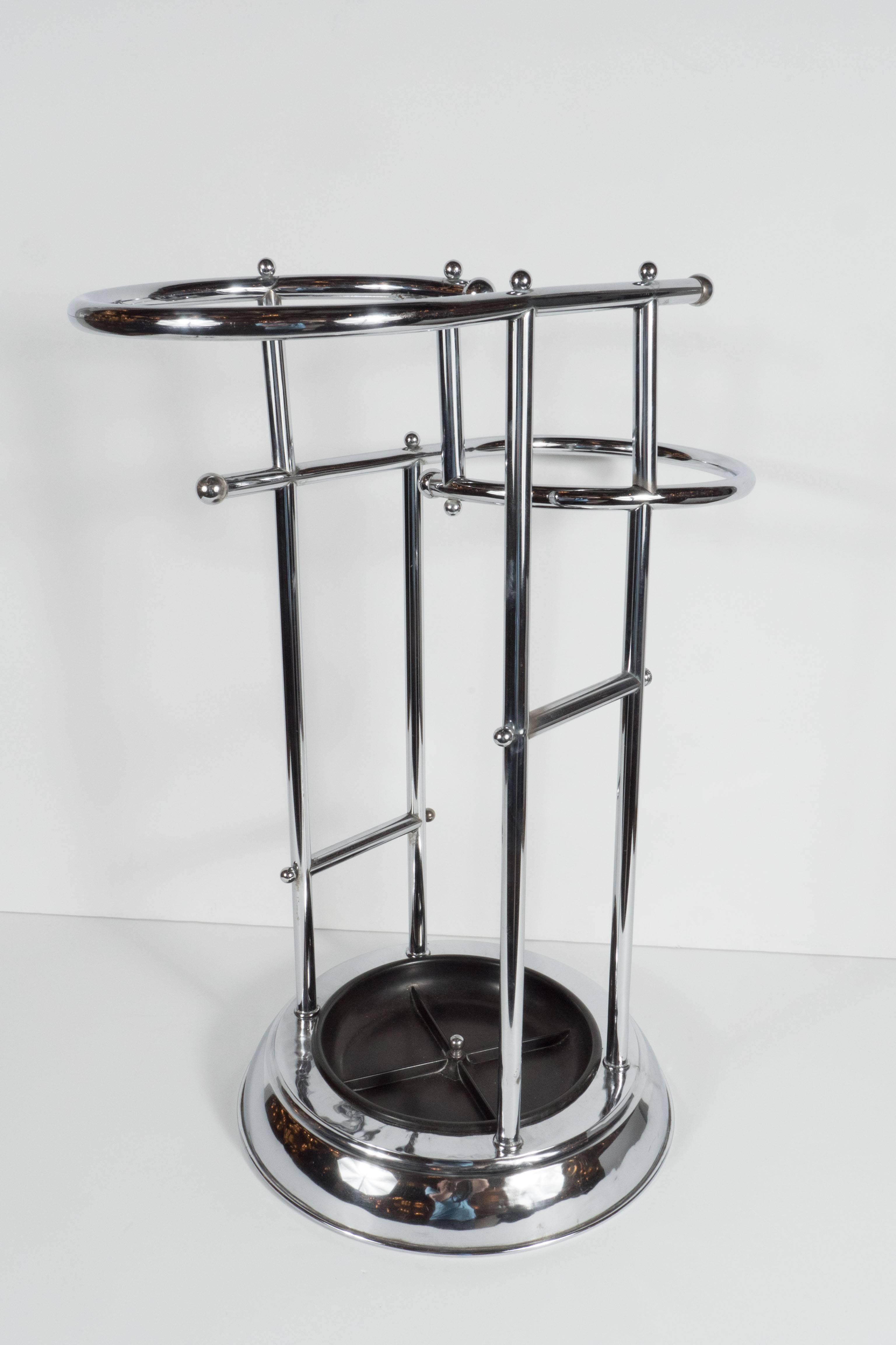 This Machine Age Art Deco umbrella stand features a stylized Bauhaus inspired design in chrome. It features a circular base with an inset bakelite center to catch dripping from wet umbrellas. The chromed tubular two tiers are designed for taller and