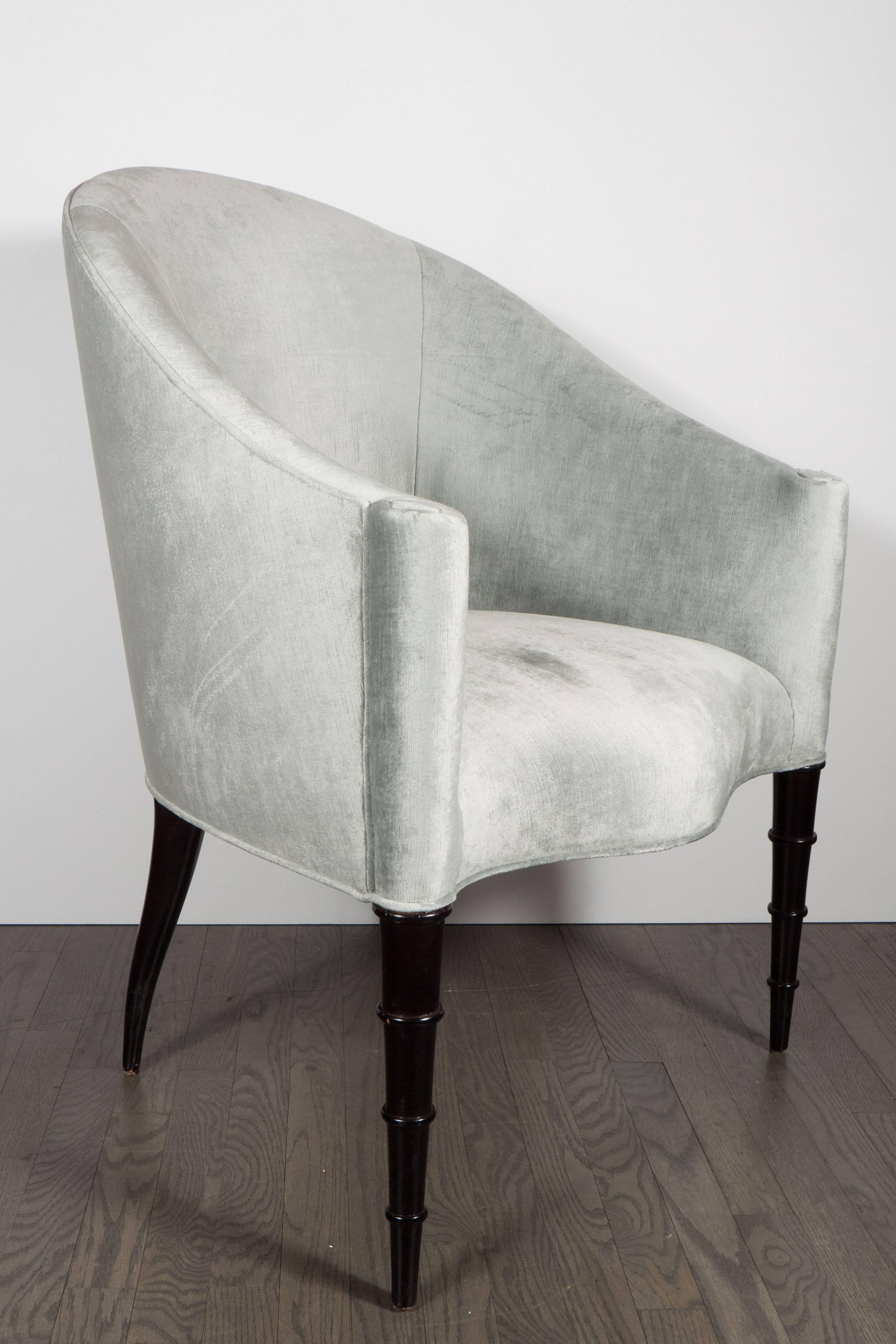 A pair of Hollywood Regency gondola chairs in pale grey platinum velvet upholstery. The legs are hand-rubbed ebonized walnut. Scroll detailing makes up the arm rests while sculptural front and splayed read legs lend to a sophisticated look. They