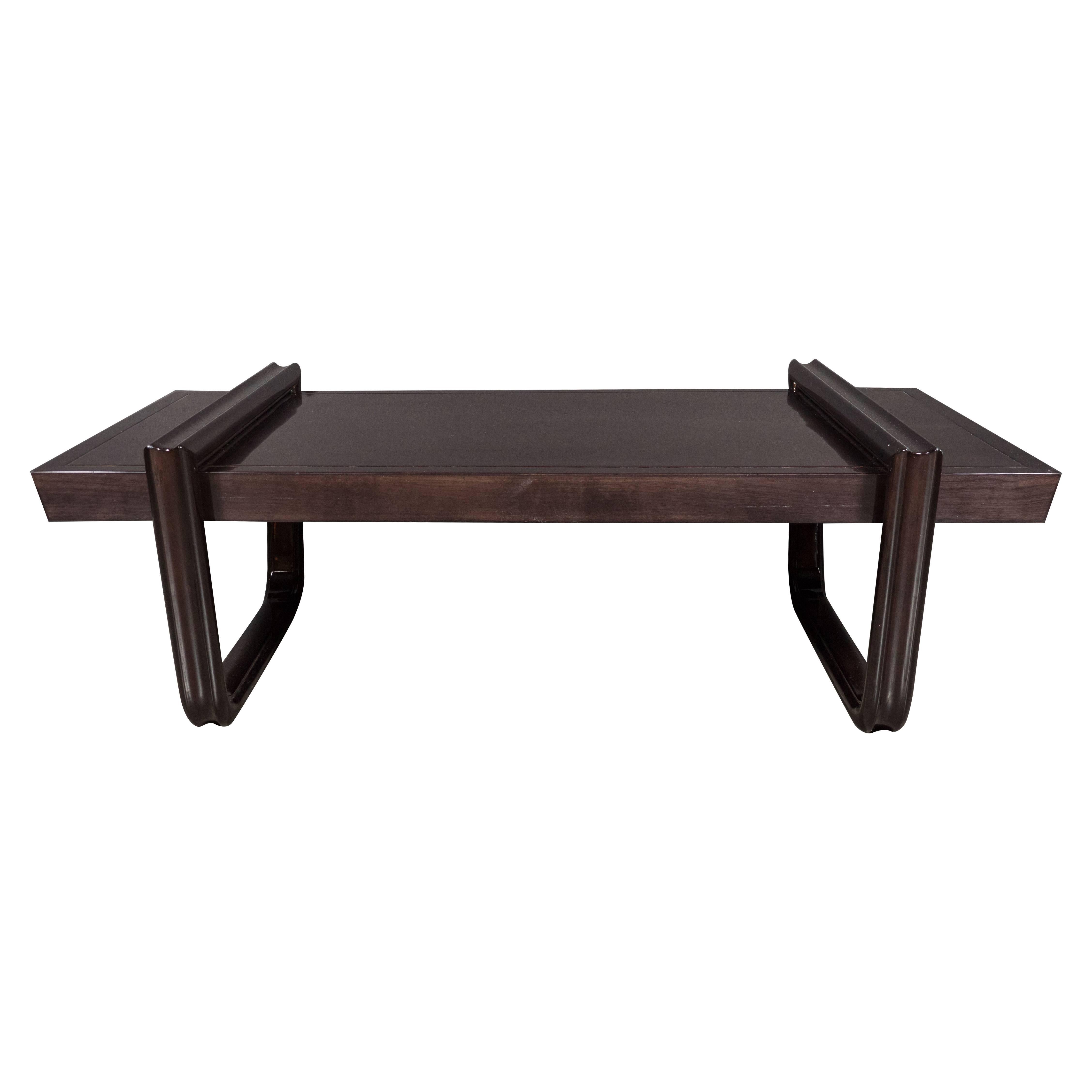 A Mid-Century cocktail or coffee table in ebonized walnut. Two continuous, wraparound legs support a rectangular table-top. Thin detailing adorns the perimeter of the top of the piece, while inward-curved legs make up the supports of the table.