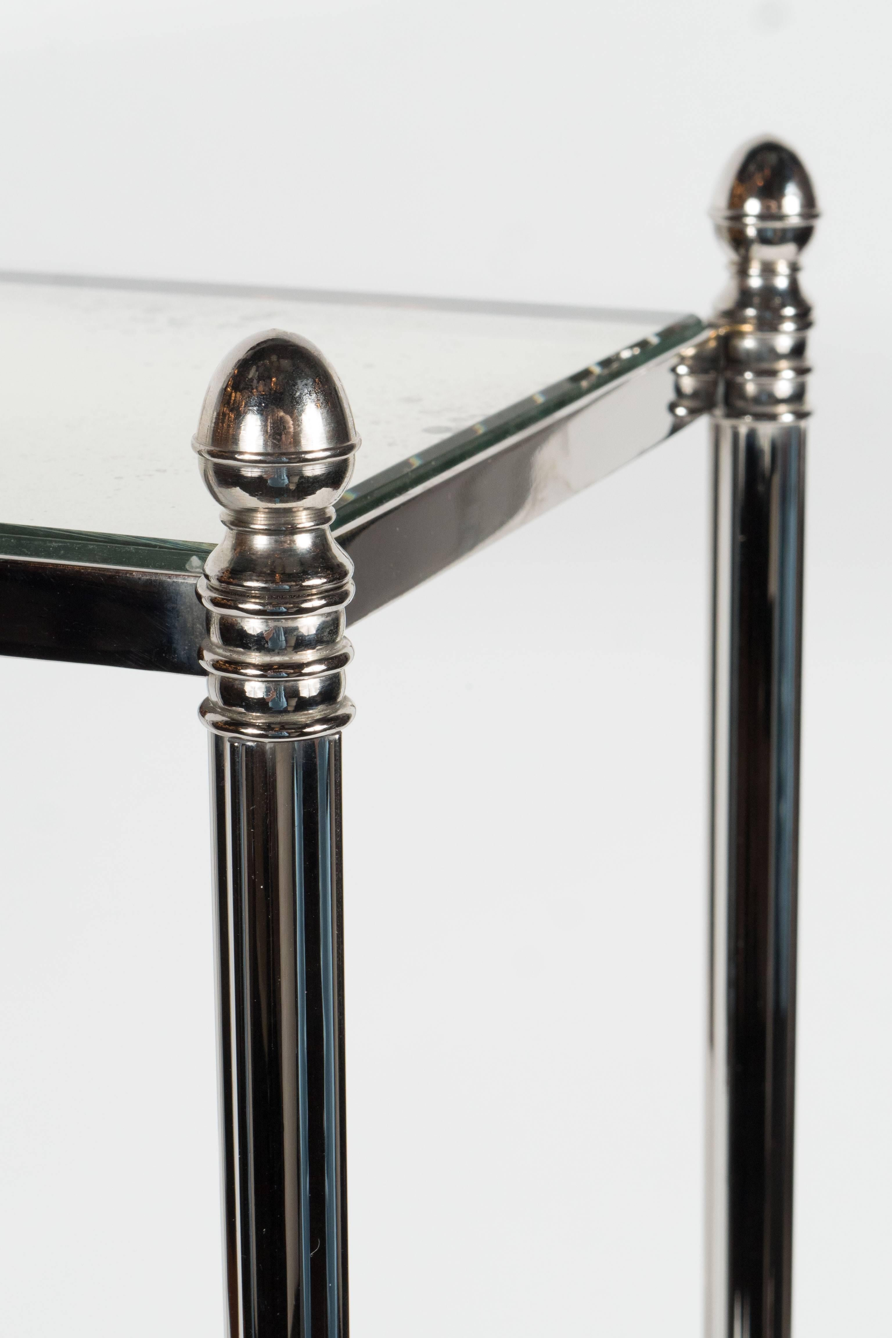 A pair of Mid-Century Modernist drinks or side tables in the manner of Jansen in polished nickel. Four legs and lower cross-bar supports hold an antiqued mirrored top with finial detailing on the four corners. An elegant pair to anchor a sofa or