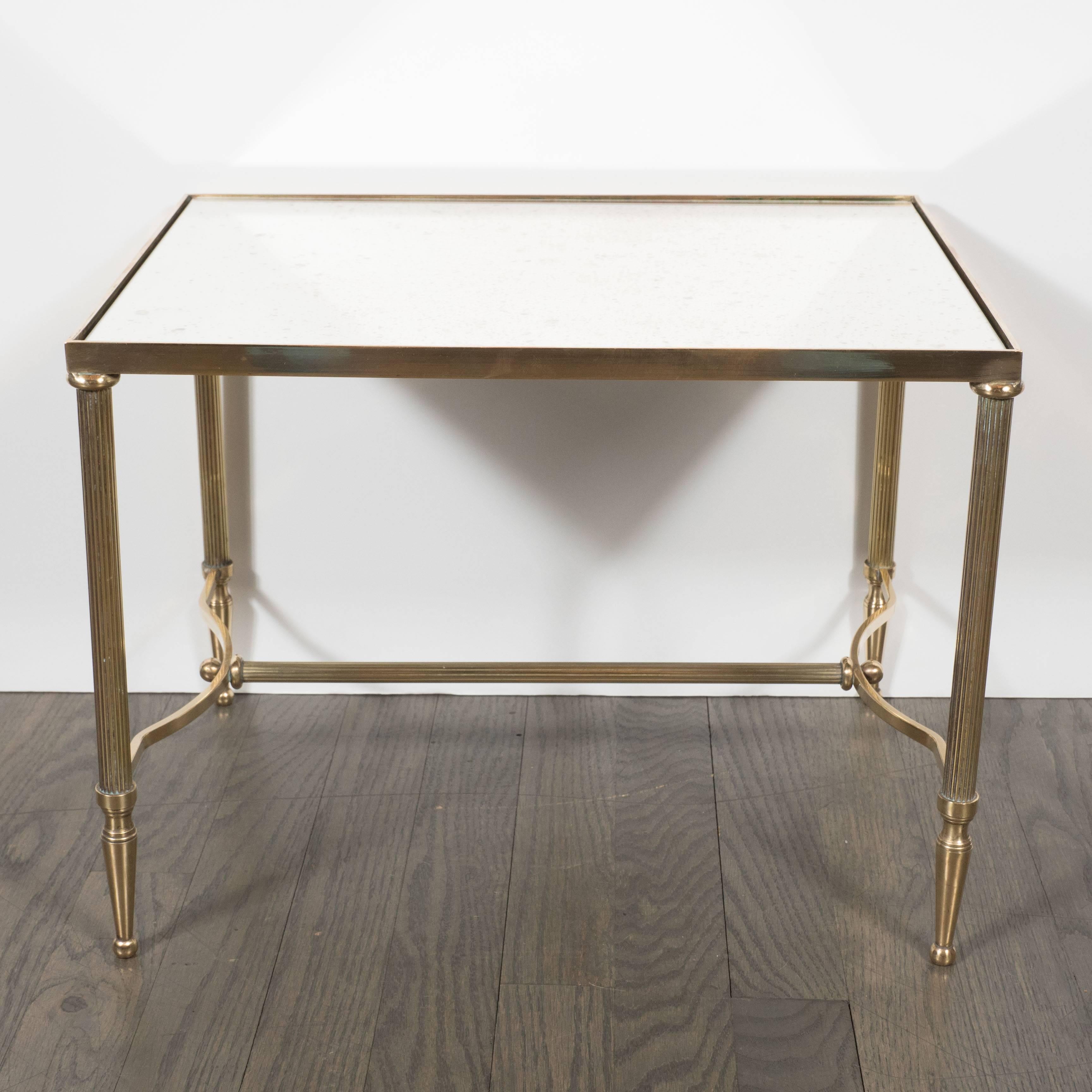 An occasional table in brass and antique mirror in the manner of Jansen. Spiraling ribbed legs are detailed with tapered feet with ball points. Cross-supports attached by a connecting rod connect opposing sides. An inset, antique mirrored top is
