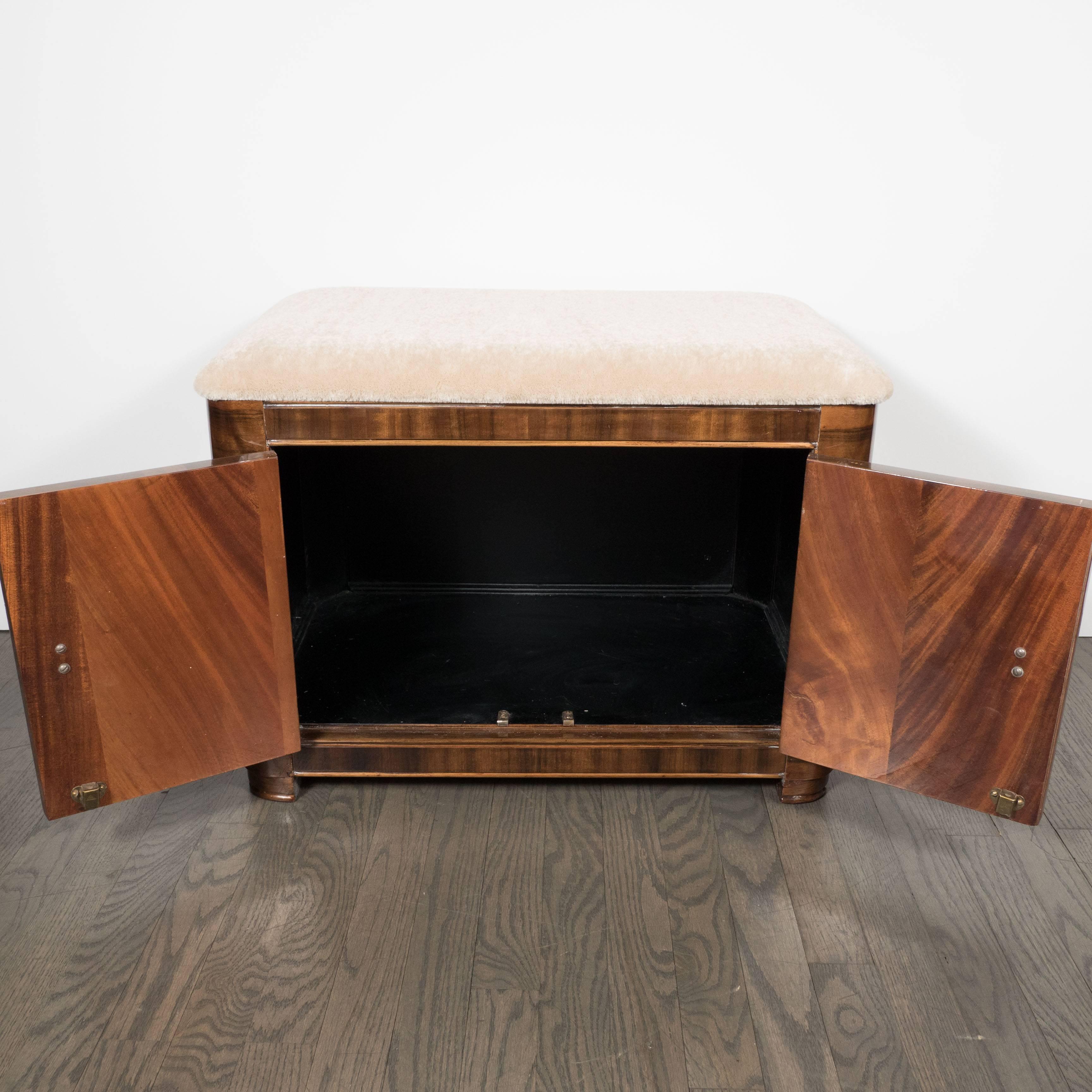An Art Deco Machine Age storage bench in bookmatched walnut with original Bakelite and brass pulls. A newly upholstered seat or foot rest in luxe camel mohair top a bookmatched base. A pair of center-handled doors open to reveal ample storage. A