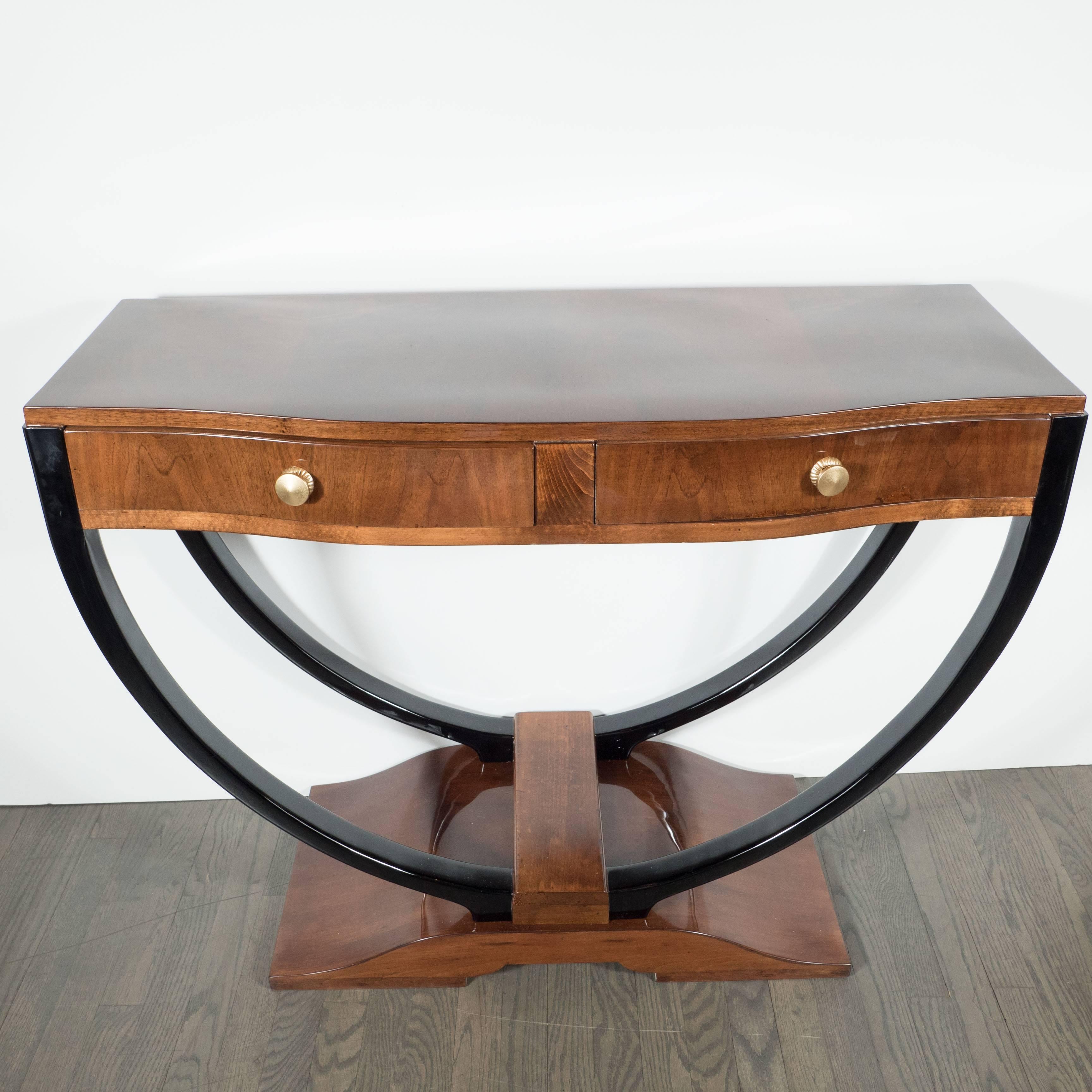 A French Art Deco console in lacquered bookmatched walnut with black lacquer accents. A curved base holds double U-form supports, on which rests a console with two pull-drawers, each adorned with a stylized round brass knob. The facade has a slight