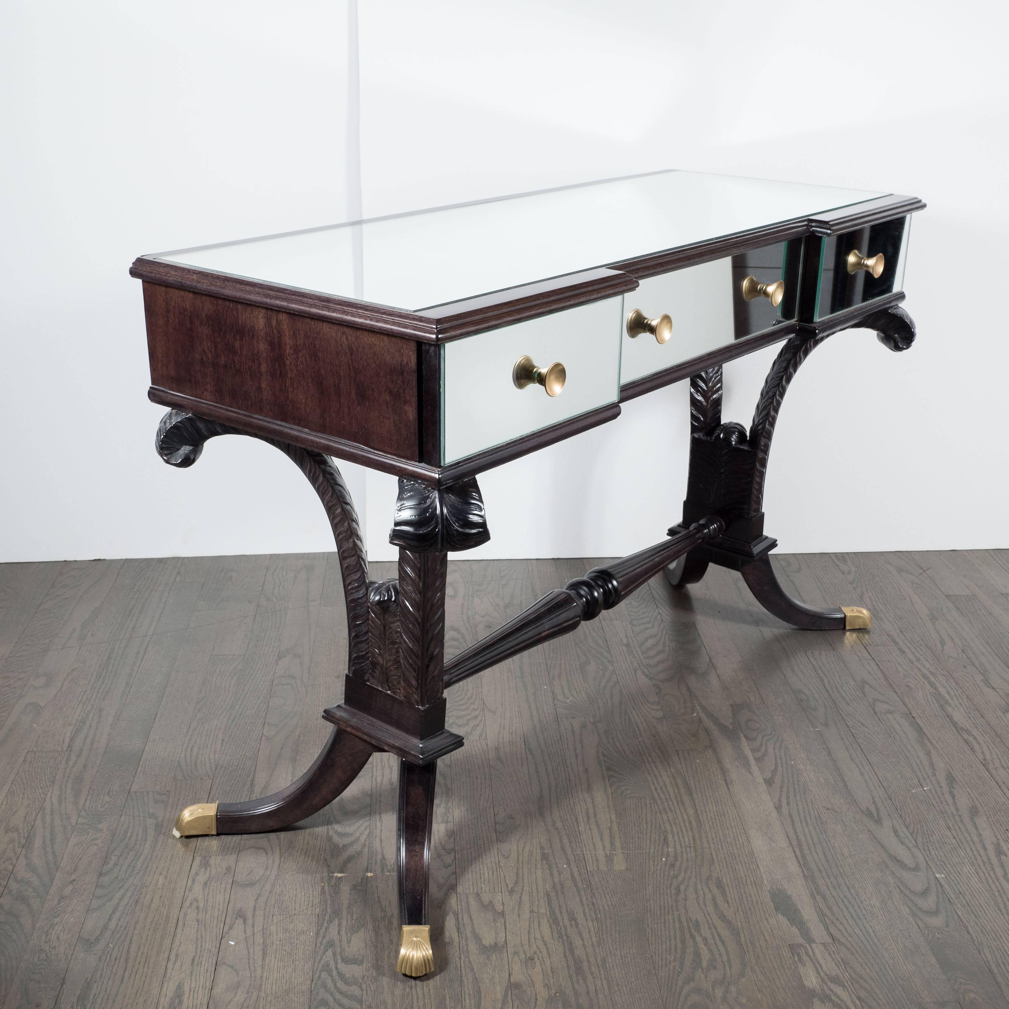 A Hollywood Regency mirrored vanity or desk by Grosfeld House in ebonized walnut and brass. A hand-rubbed ebonized walnut base consisting or four splayed legs with brass feet and a plume design support a mirrored top with drawers. A detailed