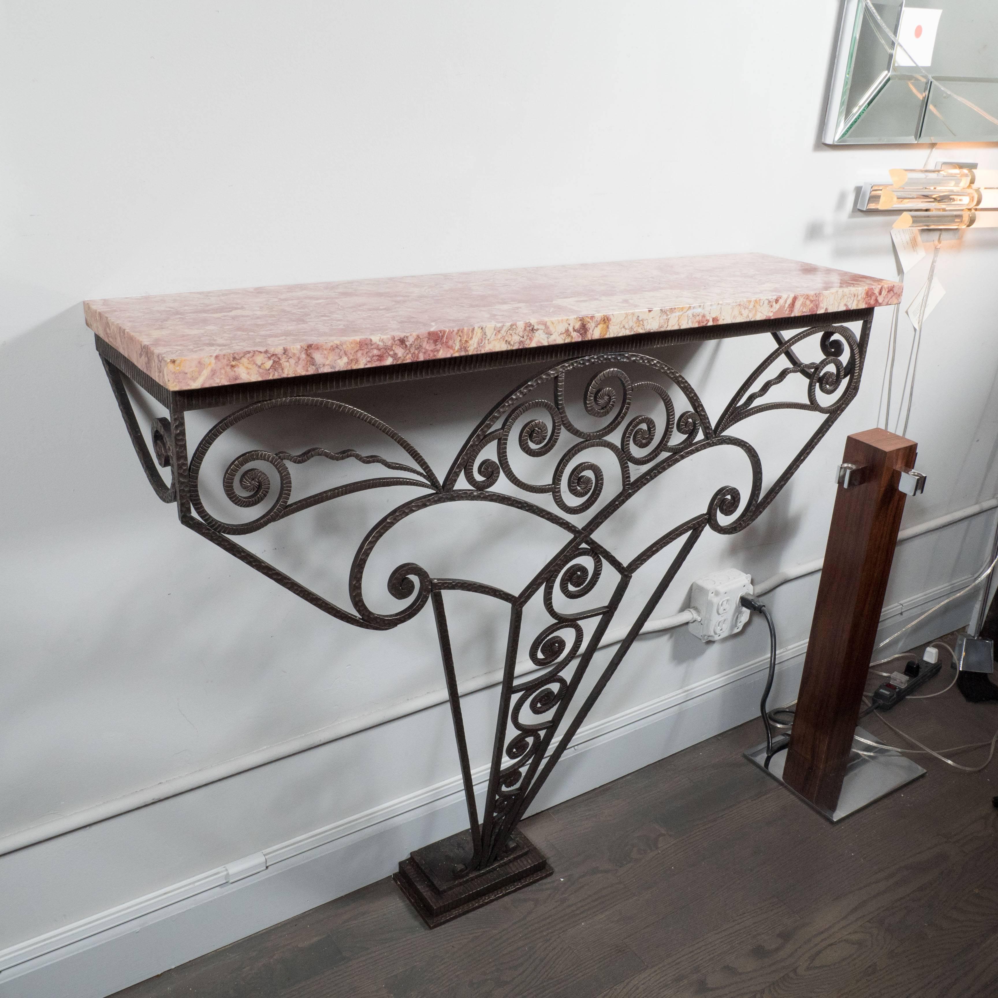 An Art Deco wall-mounted console in the manner of Edgar Brandt, the base on this console is an elaborate geometric amalgamation of wrought iron scrolls and arches branching out to support an exotic marble top in tones of fuchsia, rose and ivory. The