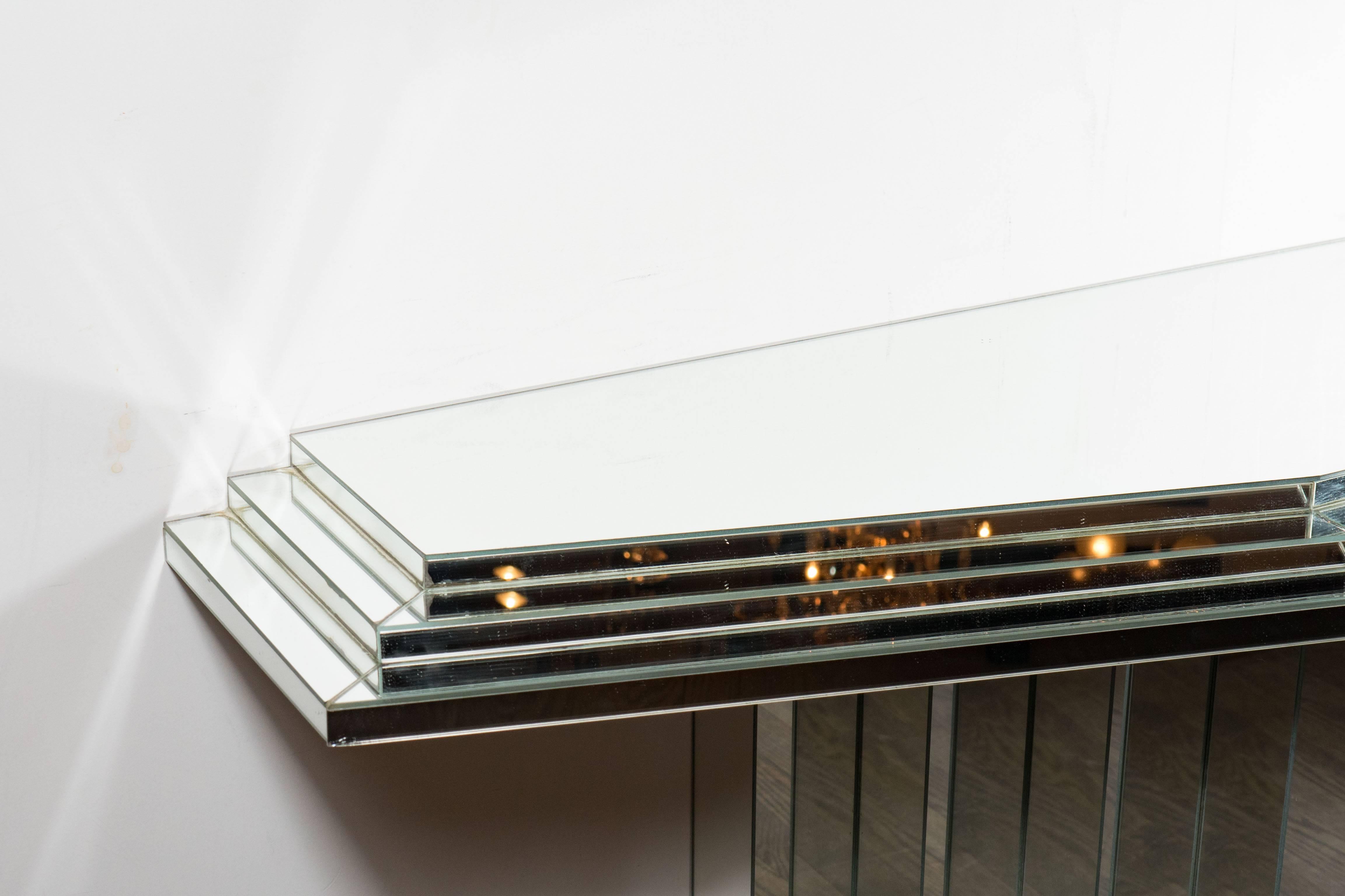 A Mid-Century Modernist mirrored console with skyscraper style detailing throughout. The top consists of three layers with the widest points tapering out to give this piece dimension. The base is also stepped spanning out from the center. It is in