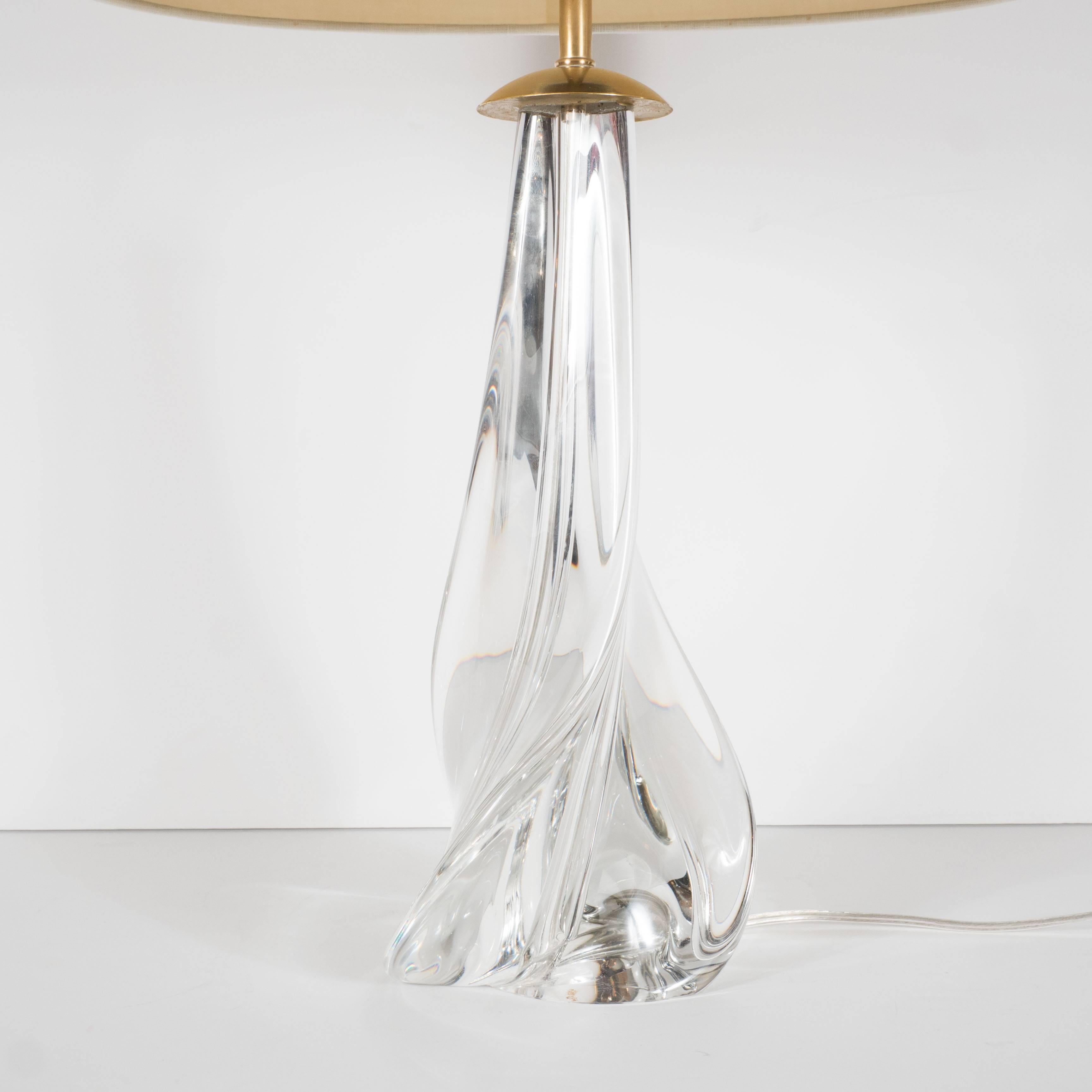 A crystal swirl-form table or desk lamp by St. Louis. A single piece of handblown crystal with a curved swirl-like form is topped by a polished brass dome-cap. A custom, oval linen shade covers two standard candelabra-based sockets. A domed,