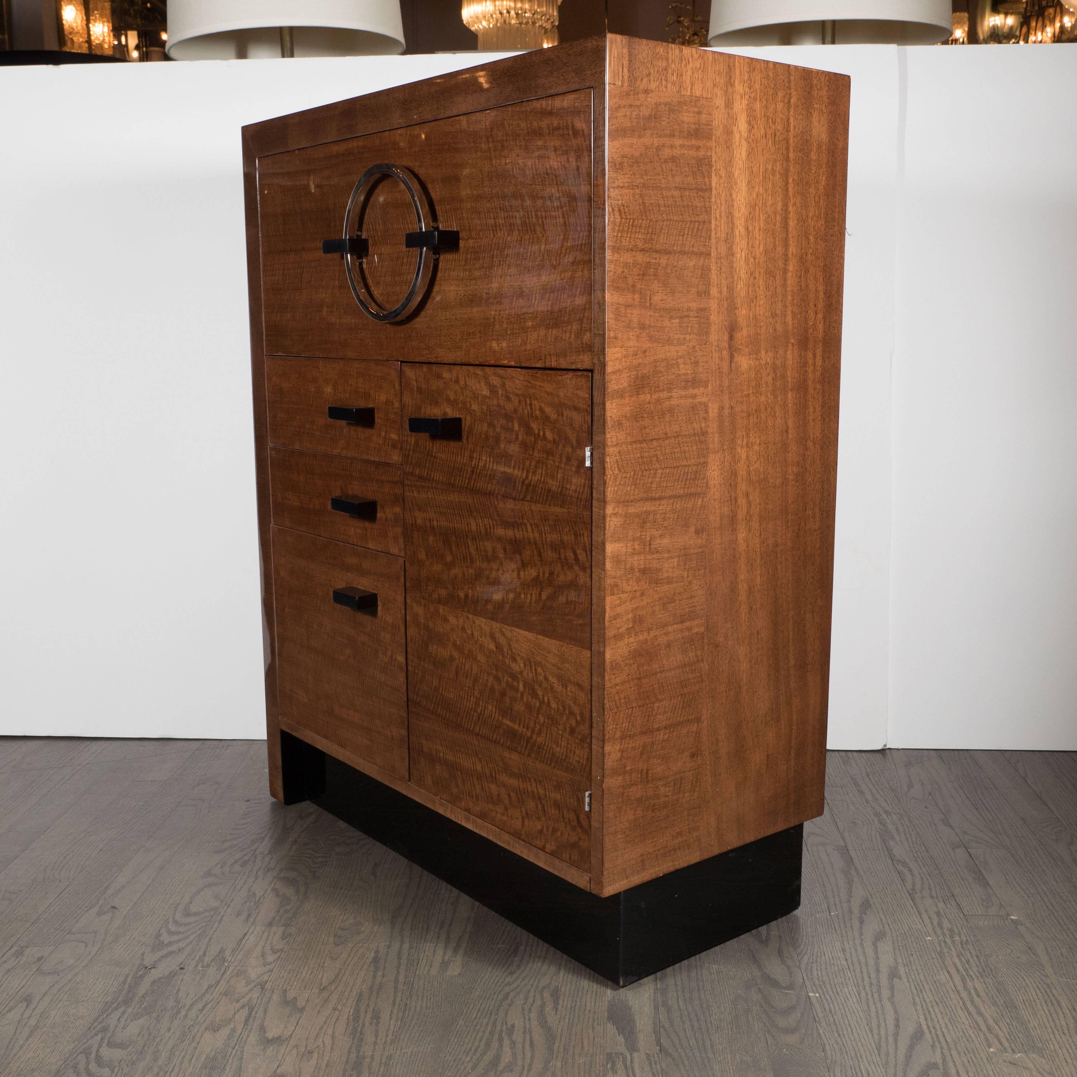An streamline Machine Age Art Deco cabinet or secretaire by Gilbert Rohde in bookmatched East Indian Laurel with black lacquer accents featuring a drop-down door which covers several compartments and a drawer. This piece has a polished nickel