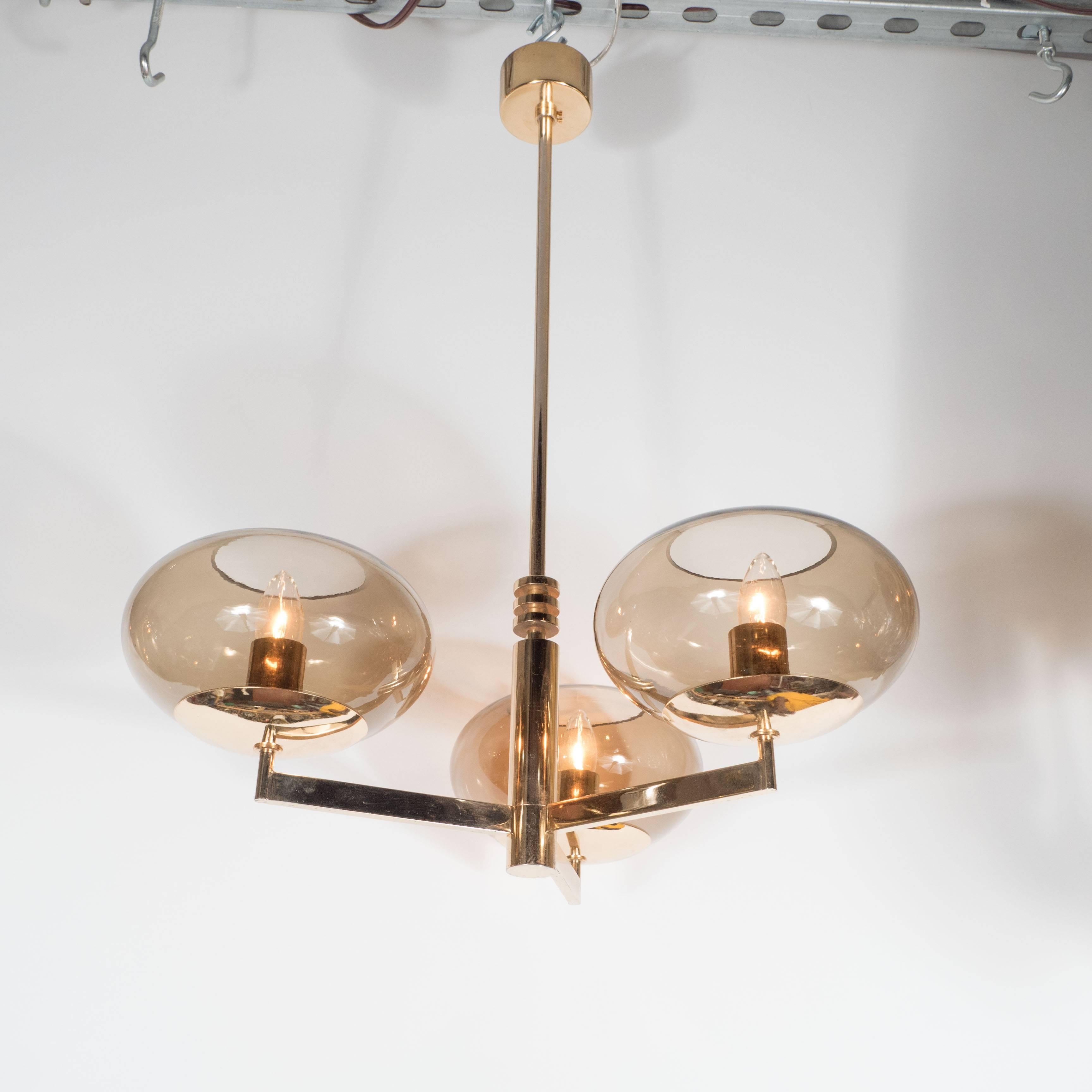 An Italian Mid-Century smoked glass and polished brass three-arm chandelier by Gaetano Sciolari. A brass central rod with disk detailing supports three arms, each fitted with an open smoked glass shade and standard candelabra socket, each capable of