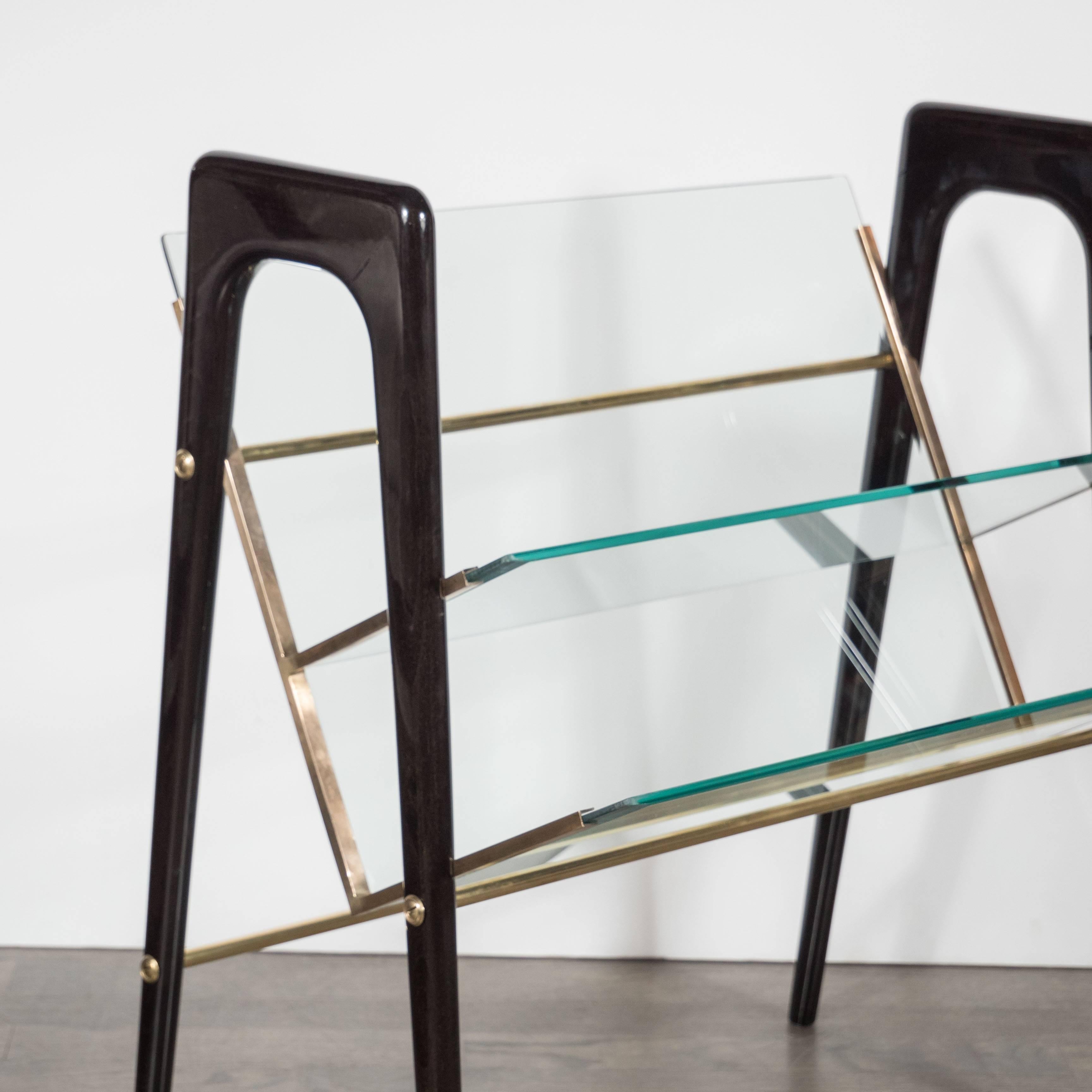 A Mid-Century modernist magazine stand in ebonized walnut, polished brass and glass in the manner of Ico Parisi. Round curved edges support two separate glass shelves which can hold magazines or books. A brass frame supports the shelves. This piece