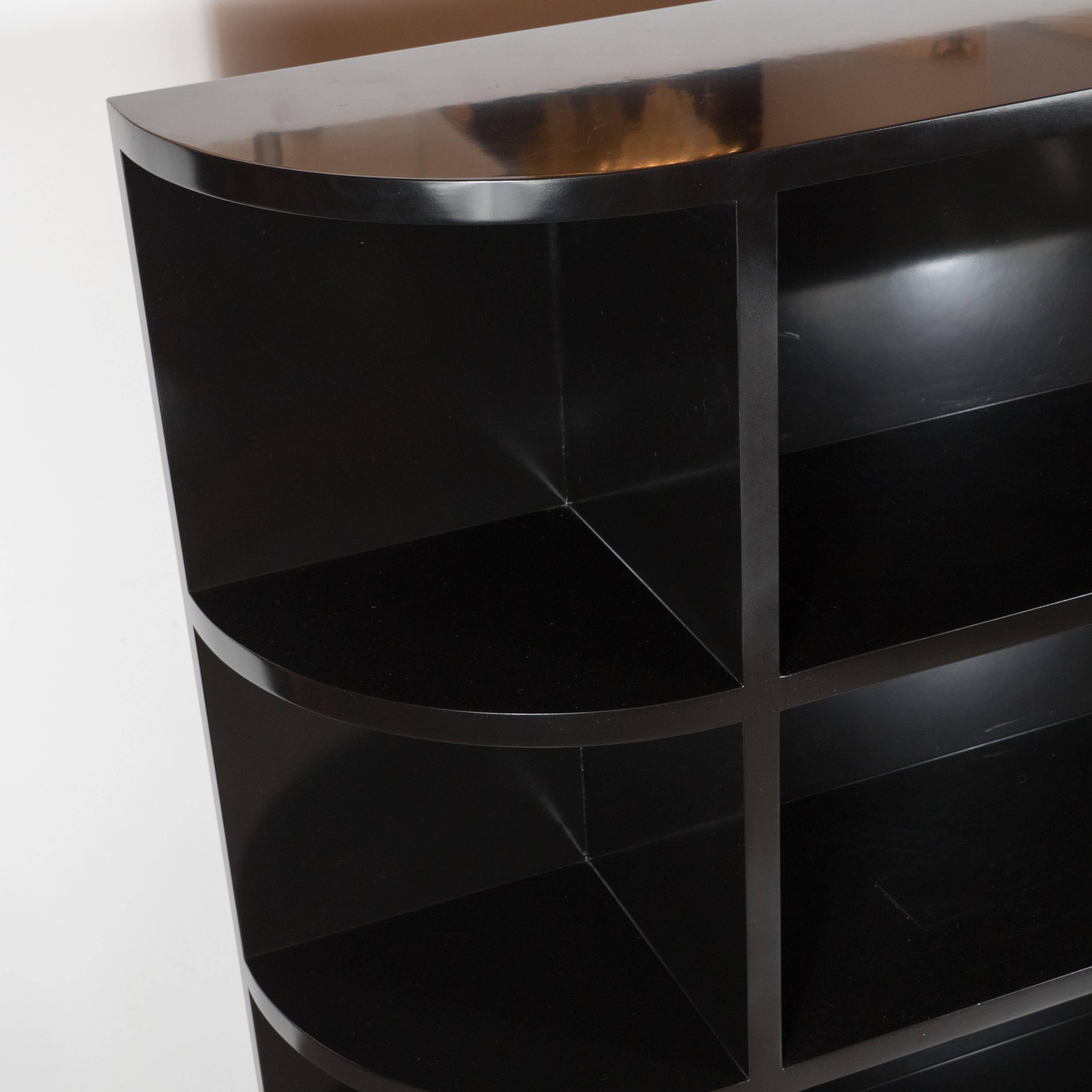 
A striking Streamline Art Deco black lacquered chest level bookcase with one straight end and the other semi-circular end, complete with two book shelves measuring the length of the unit and smaller semi-circular shelves ideal for decorative