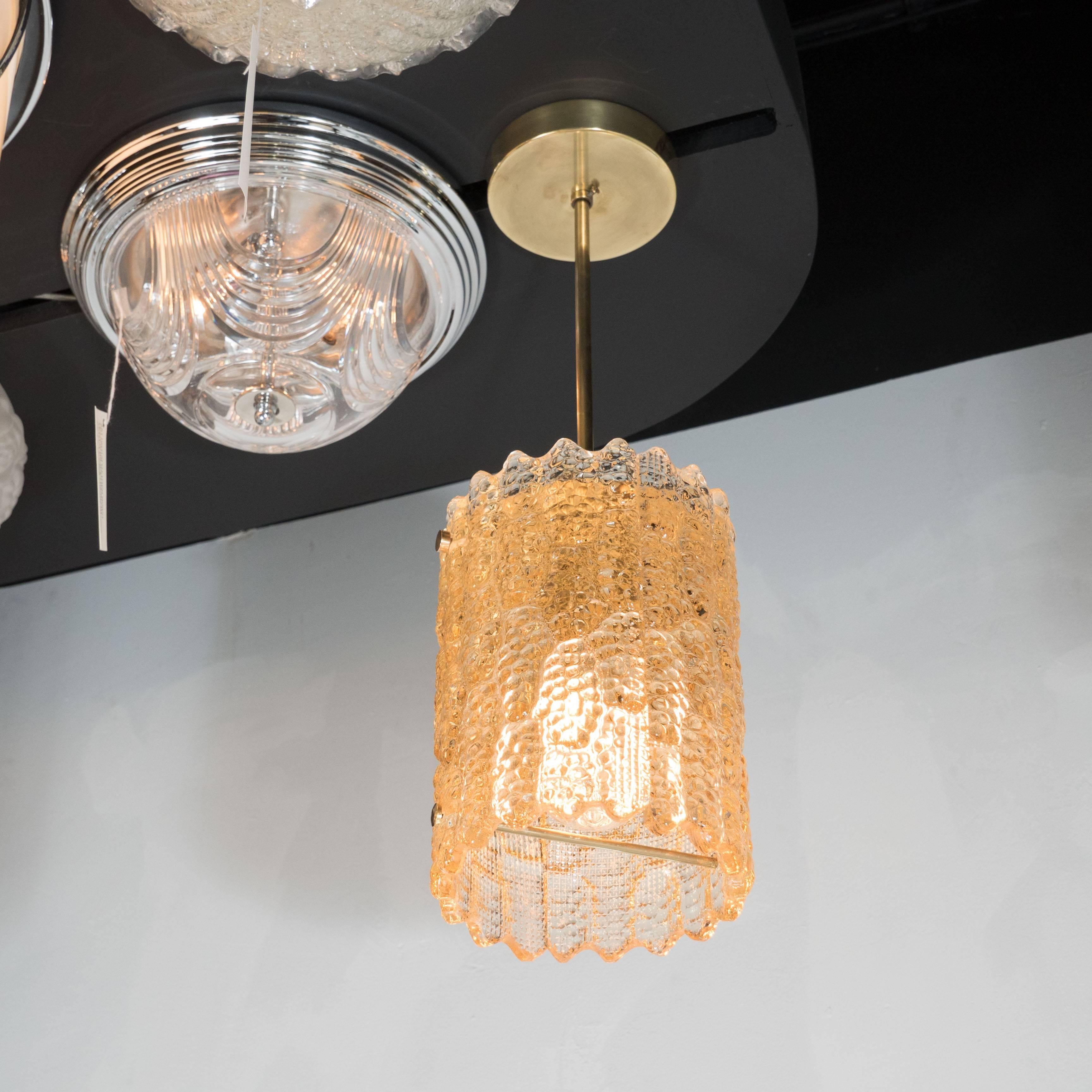 An elegant Mid-Century Modernist textured amber glass pendant with polished brass fittings by Carl Fagerlund for Orrefors of Sweden. A polished brass canopy holds a rod which supports a cylindrical form glass shade with organic textural elements and