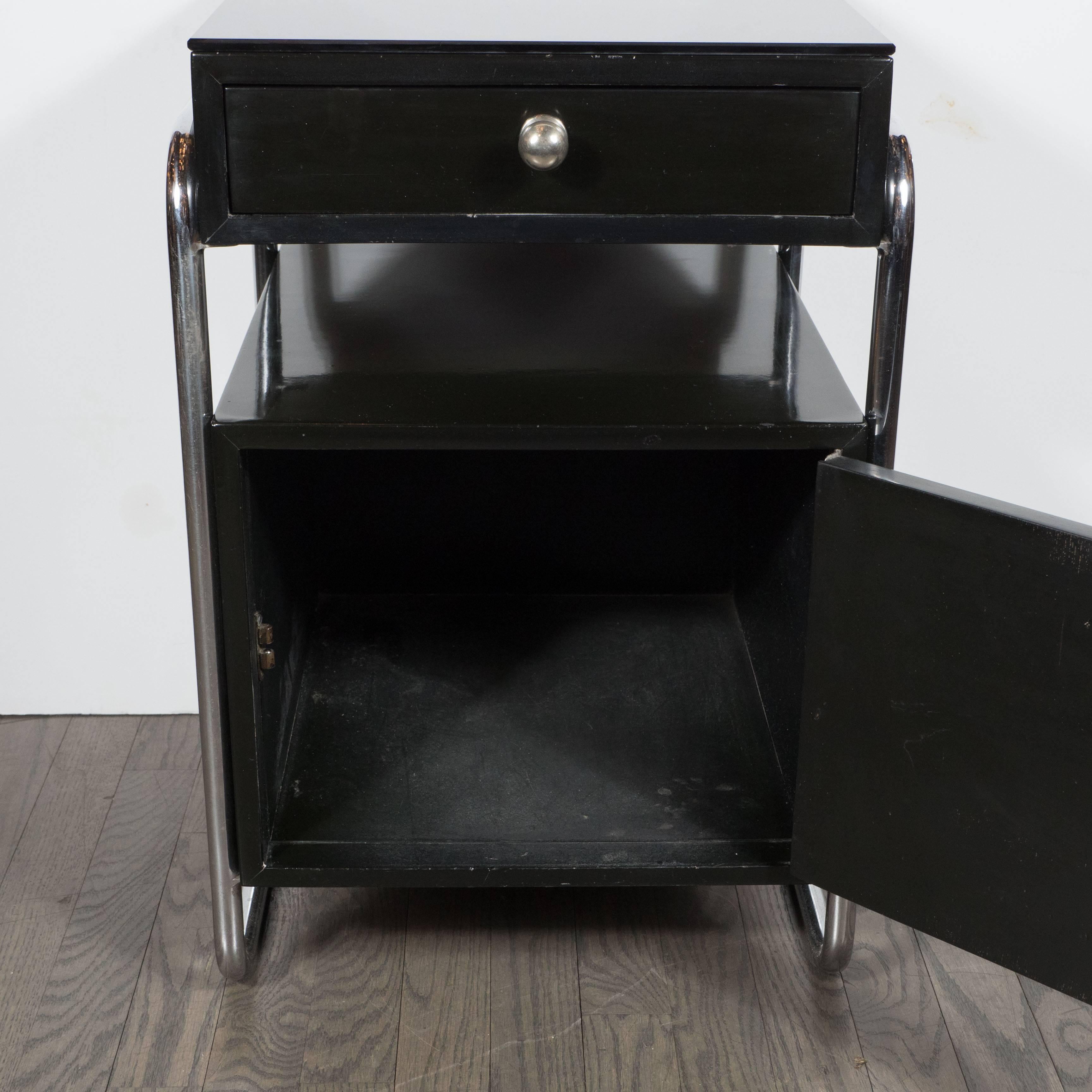 A pair of Art Deco Machine Age end tables or nightstands in black lacquer and vitrolite by Wolfgang Hoffman. Tubular chrome frames support a lower storage section with door, center shelf, and upper drawer. Chrome ball handles adorn both storage